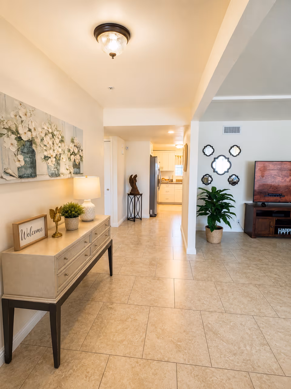 Bright hallway and living area of a senior facility with console table, plants, and TV.