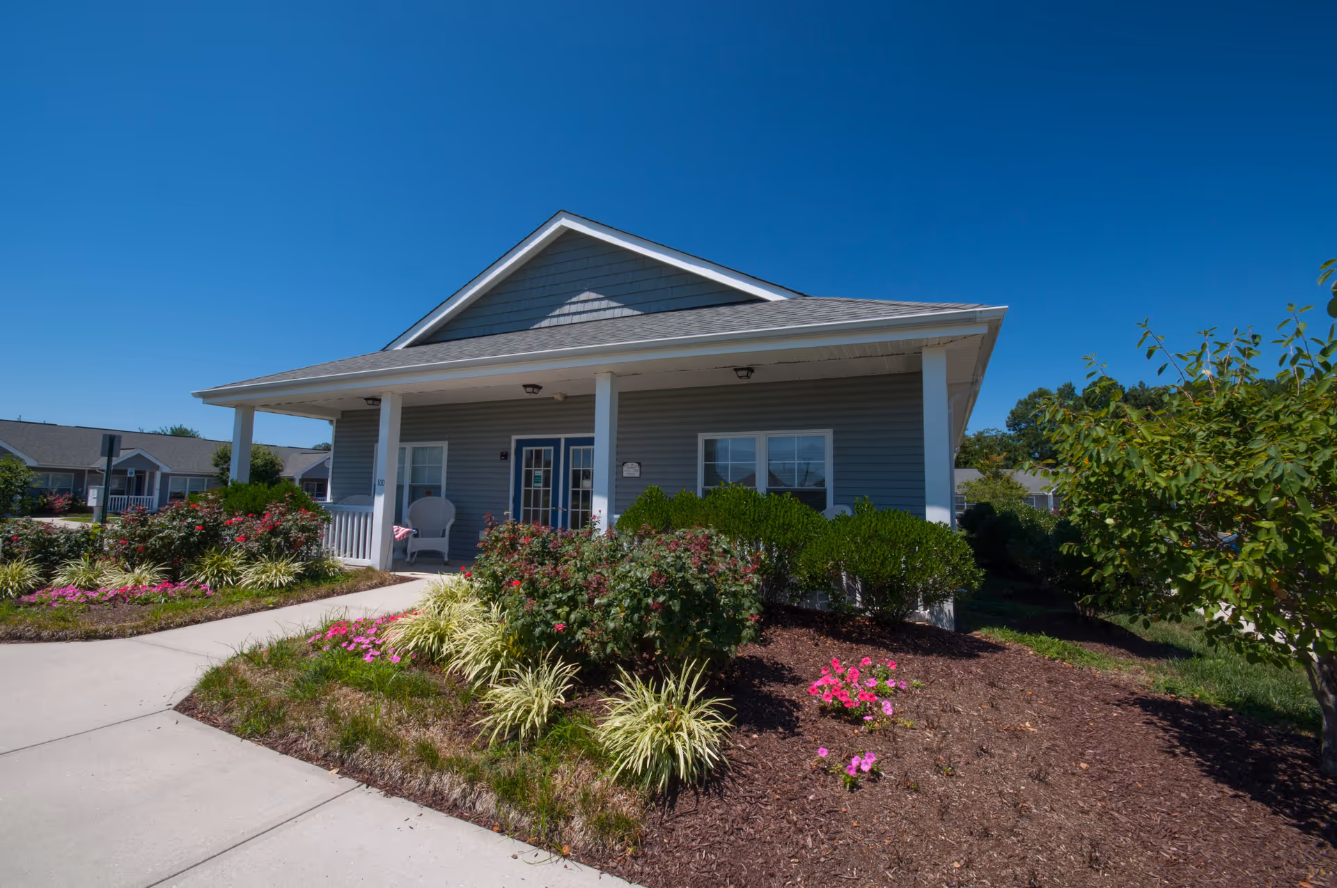 Exterior view of a single-story residential building with a covered porch, white columns, and a blue-gray siding. The porch has a white chair and a small table with a cloth. The building is surrounded by landscaped flower beds with green shrubs and pink flowers under a clear blue sky.