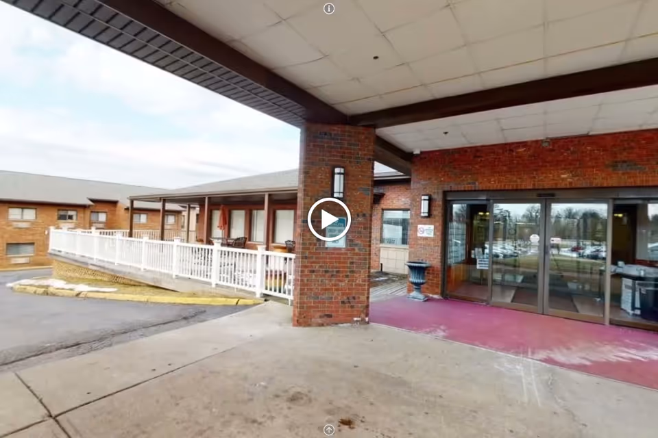 Entrance area of a senior living facility with a covered drop-off zone, brick walls, glass sliding doors, and a white railing along a ramp leading to the entrance. There are chairs and tables visible on the porch area.