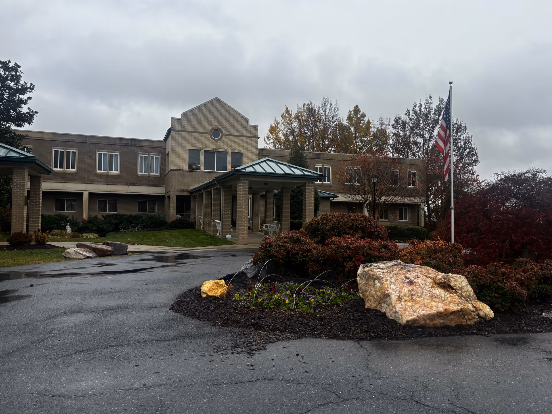 Exterior view of a two-story brick building with a covered entrance and a circular driveway. There are landscaped bushes, large rocks, and an American flag on a flagpole in front of the building under a cloudy sky.