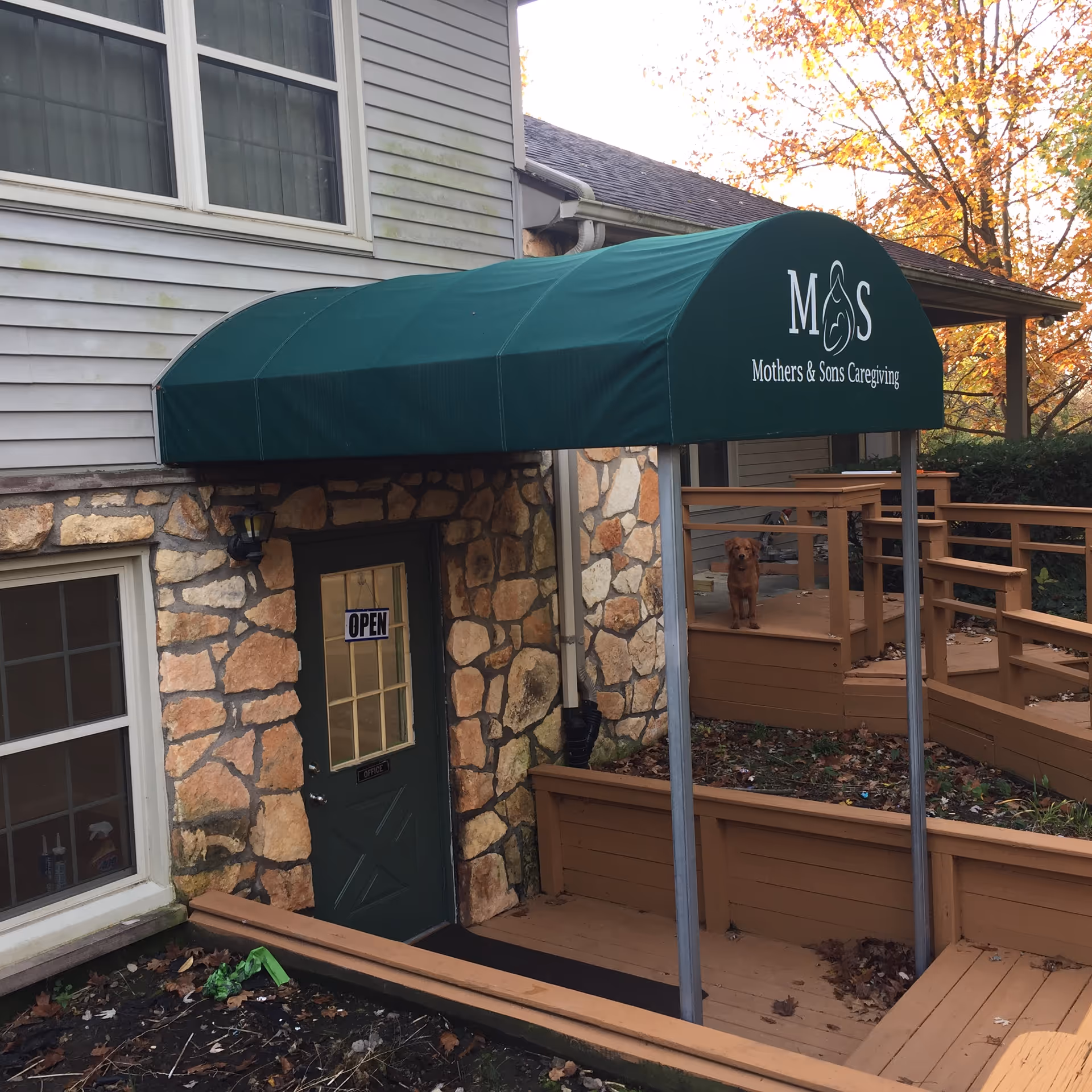 Stone building entrance with a green canopy reading Mothers & Sons Caregiving over a wooden deck.