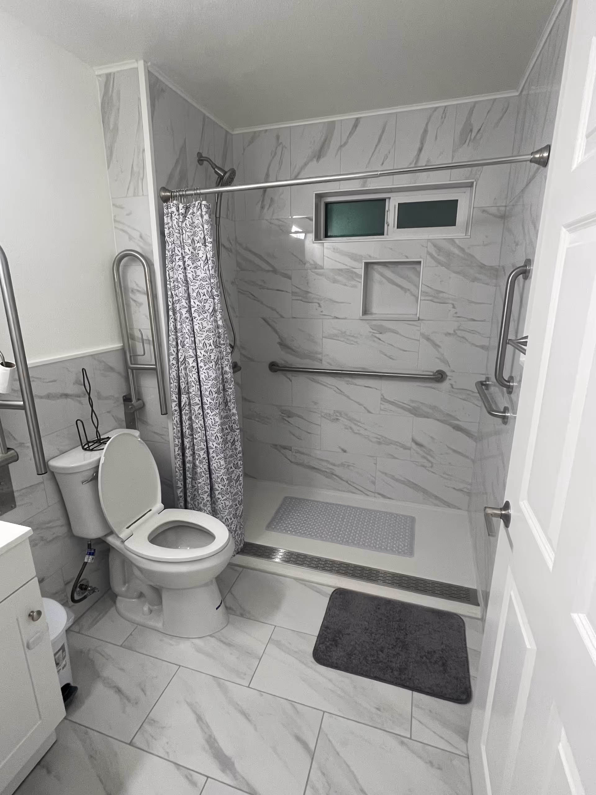 Marble tiled bathroom with toilet, shower, curtain, and multiple grab bars for accessibility.