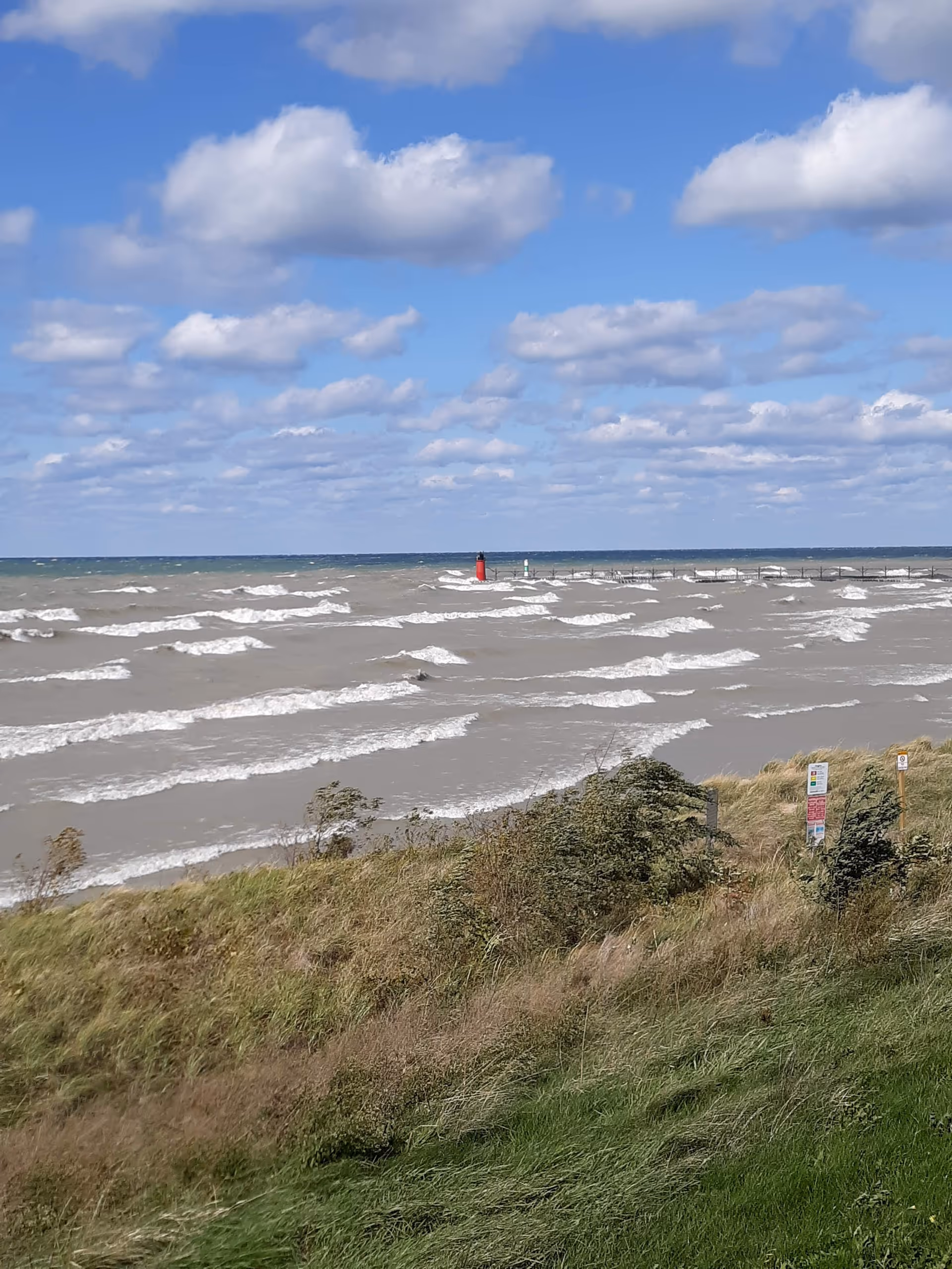 Beach scene with blue sky, white clouds, waves, and grassy dunes in the foreground.