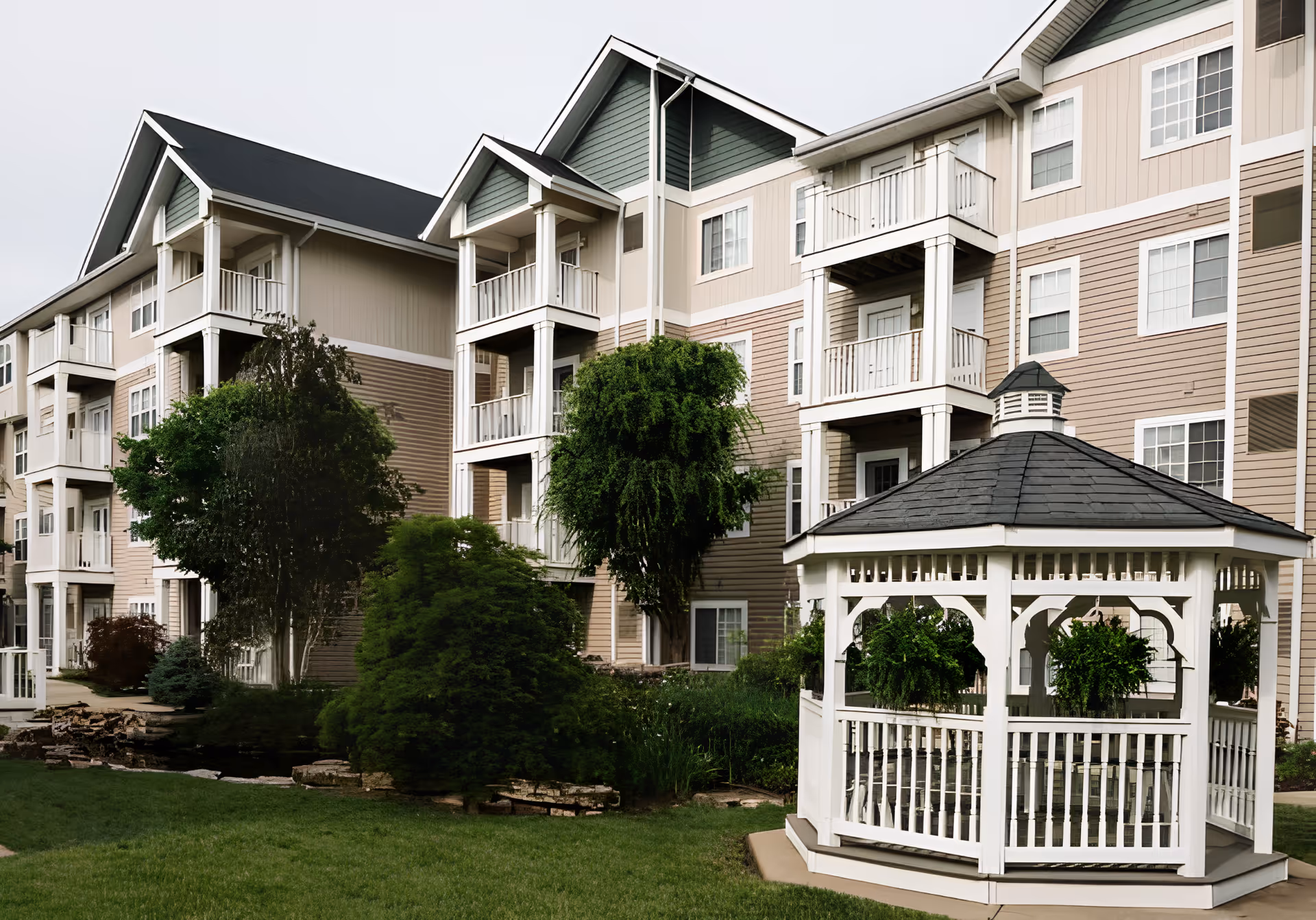 Exterior view of a multi-story senior living building with balconies, landscaped grounds, and a white gazebo in the foreground.