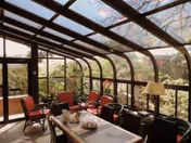 Sunlit glass-enclosed sunroom with a table and several chairs overlooking a garden.