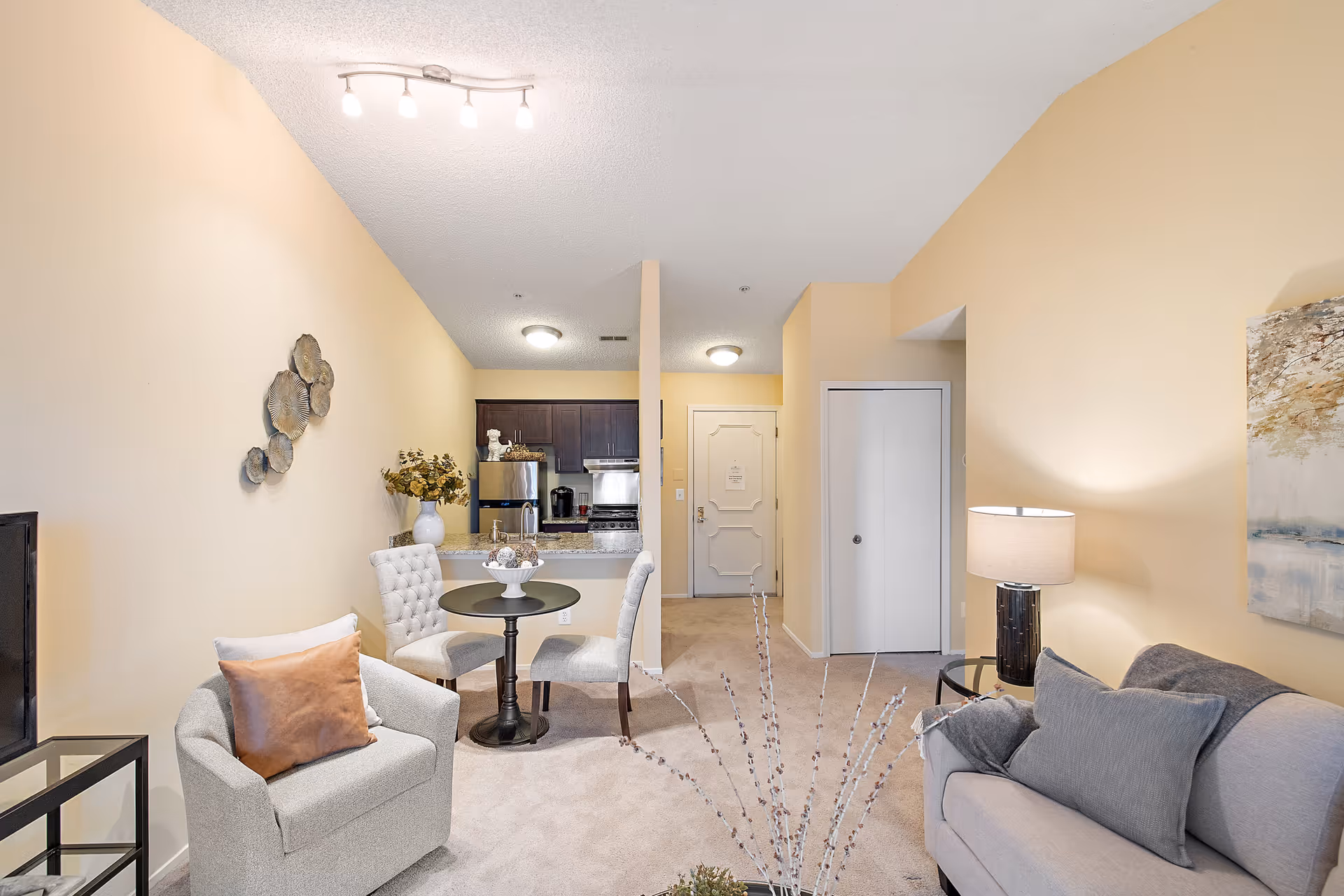 A cozy living room area with beige walls and carpet. The room features a light gray armchair with a brown pillow, a small round dining table with two upholstered chairs, a gray sofa with matching pillows, a side table with a lamp, and a wall-mounted TV. In the background, there is a kitchen with dark cabinets, stainless steel appliances, and a granite countertop. Decorative wall art and a vase with flowers add to the room's decor.