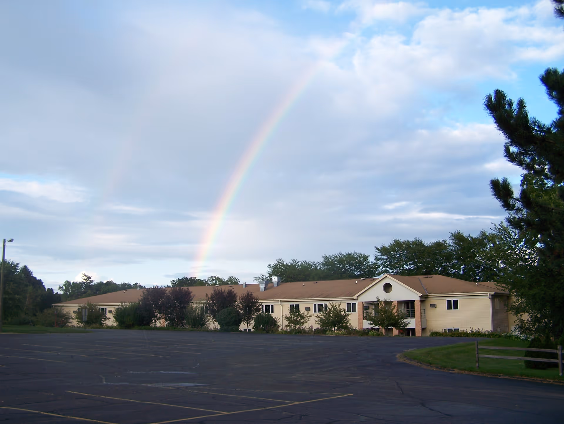 Single-story retirement center building with a rainbow in the cloudy sky and a parking lot.