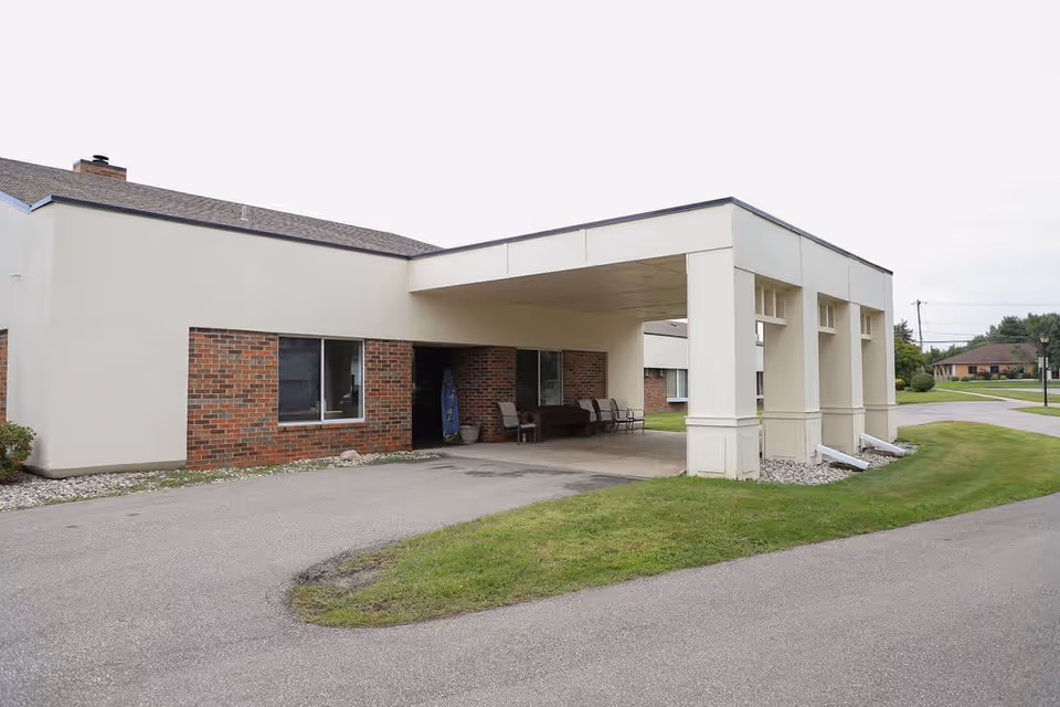 Exterior view of a single-story assisted living facility building with a covered entrance supported by large square columns. The building has a combination of brick and light-colored siding, with a paved driveway and green lawn surrounding it. Several chairs and a bench are placed under the covered entrance area.