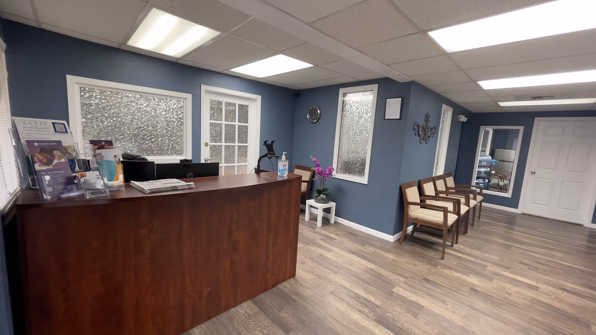 Reception desk in a blue-walled senior living lobby with chairs, windows, and a plant.