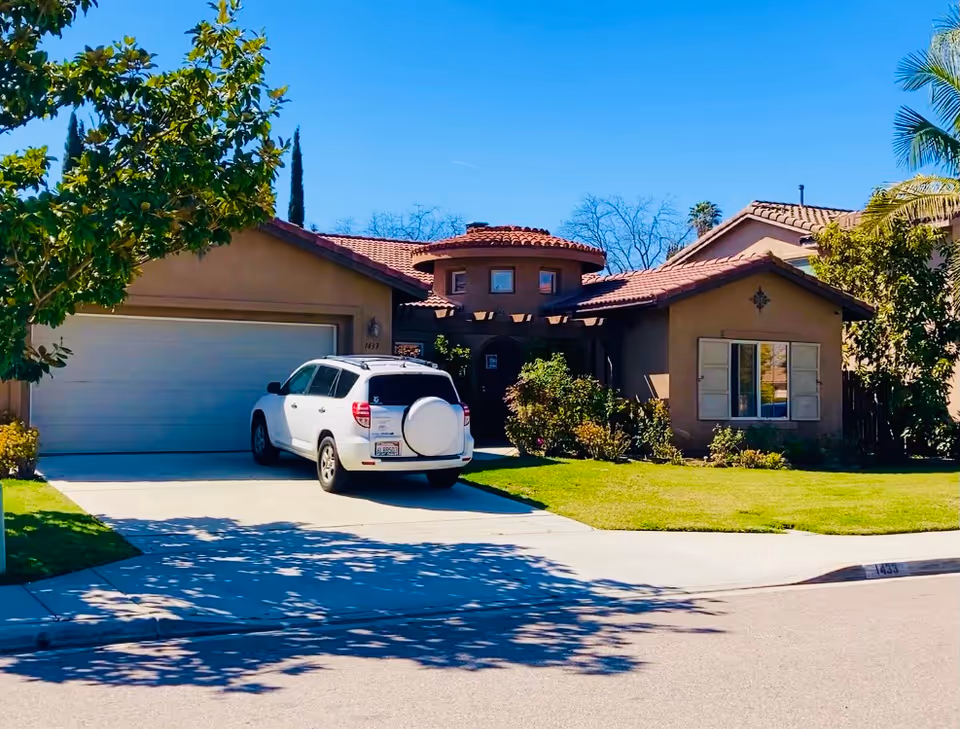 A single-story residential building with a tiled roof and a two-car garage. A white SUV is parked in the driveway. The front yard has green grass, bushes, and trees. The sky is clear and blue.