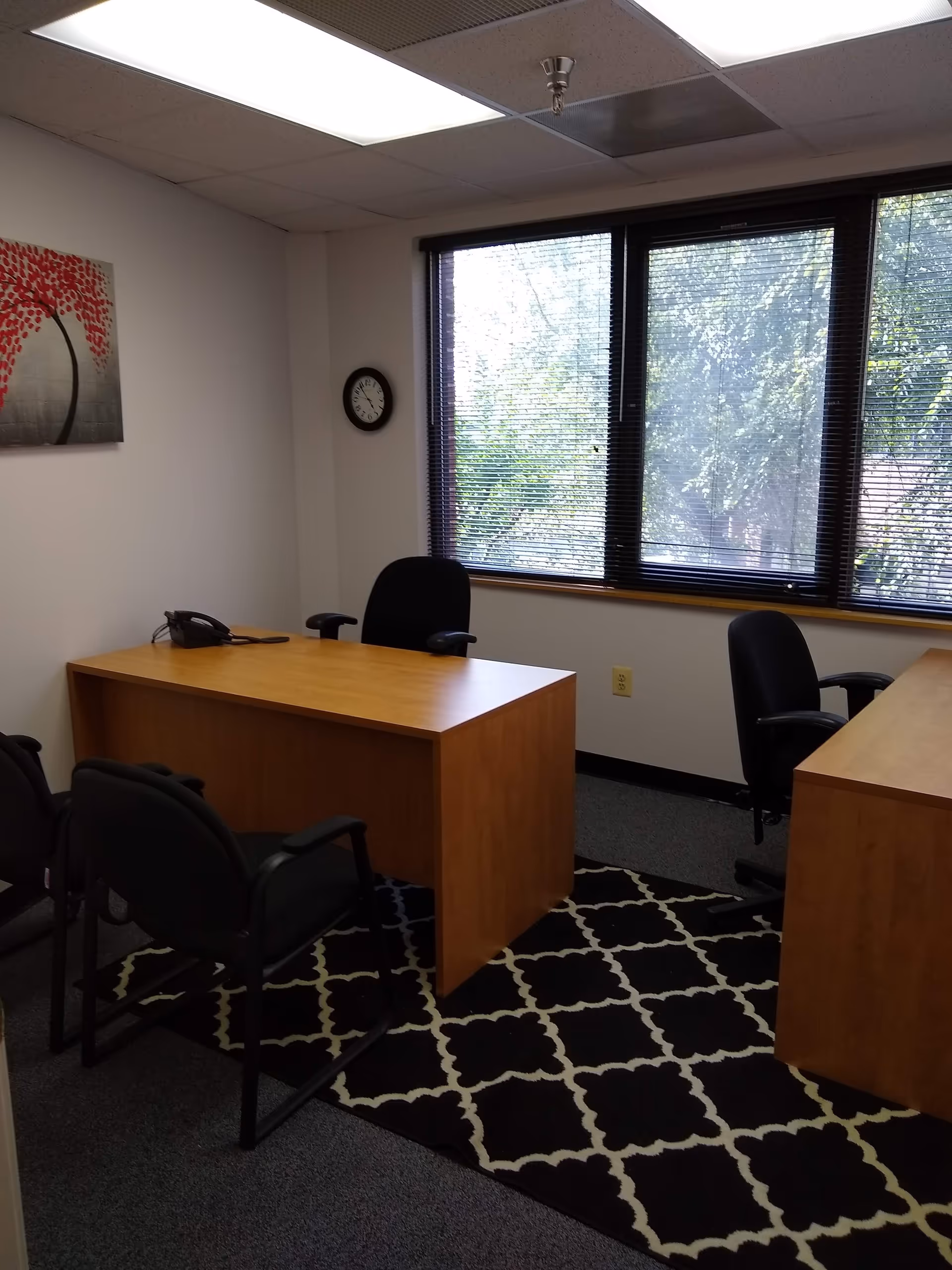 Office area with wooden desks, black chairs, blinds, rug, and wall clock.