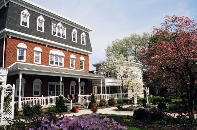 Front exterior of a multi-story brick senior living building with a covered porch and landscaped flowering trees and shrubs.