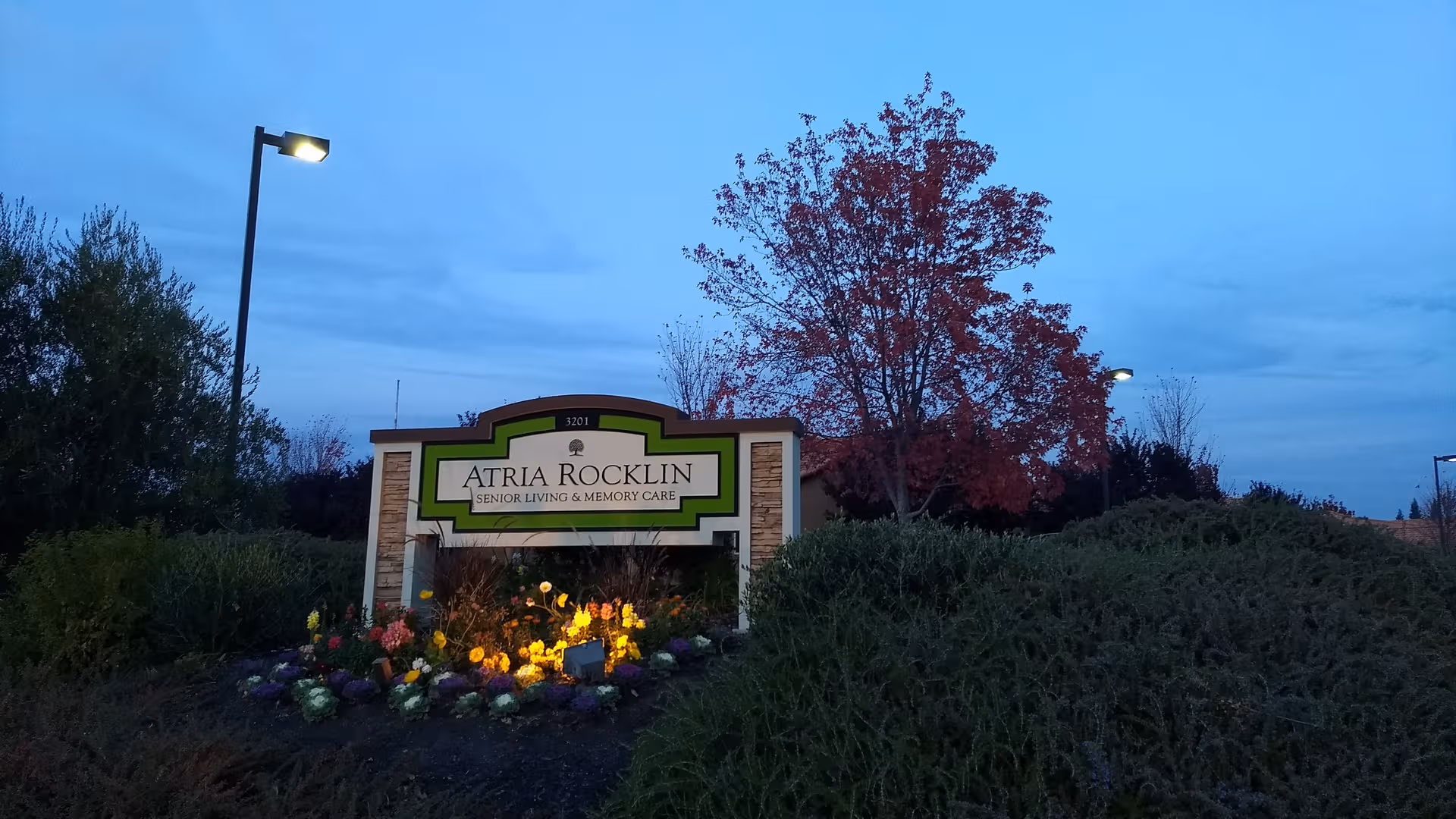 Exterior view of Atria Rocklin sign surrounded by flowers and dusk lighting.