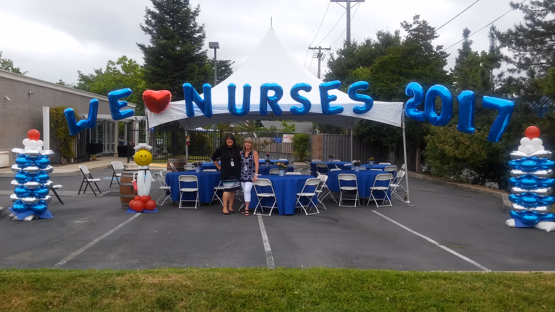 Two people stand under a white canopy with blue tablecloths and balloon decorations in a lot.