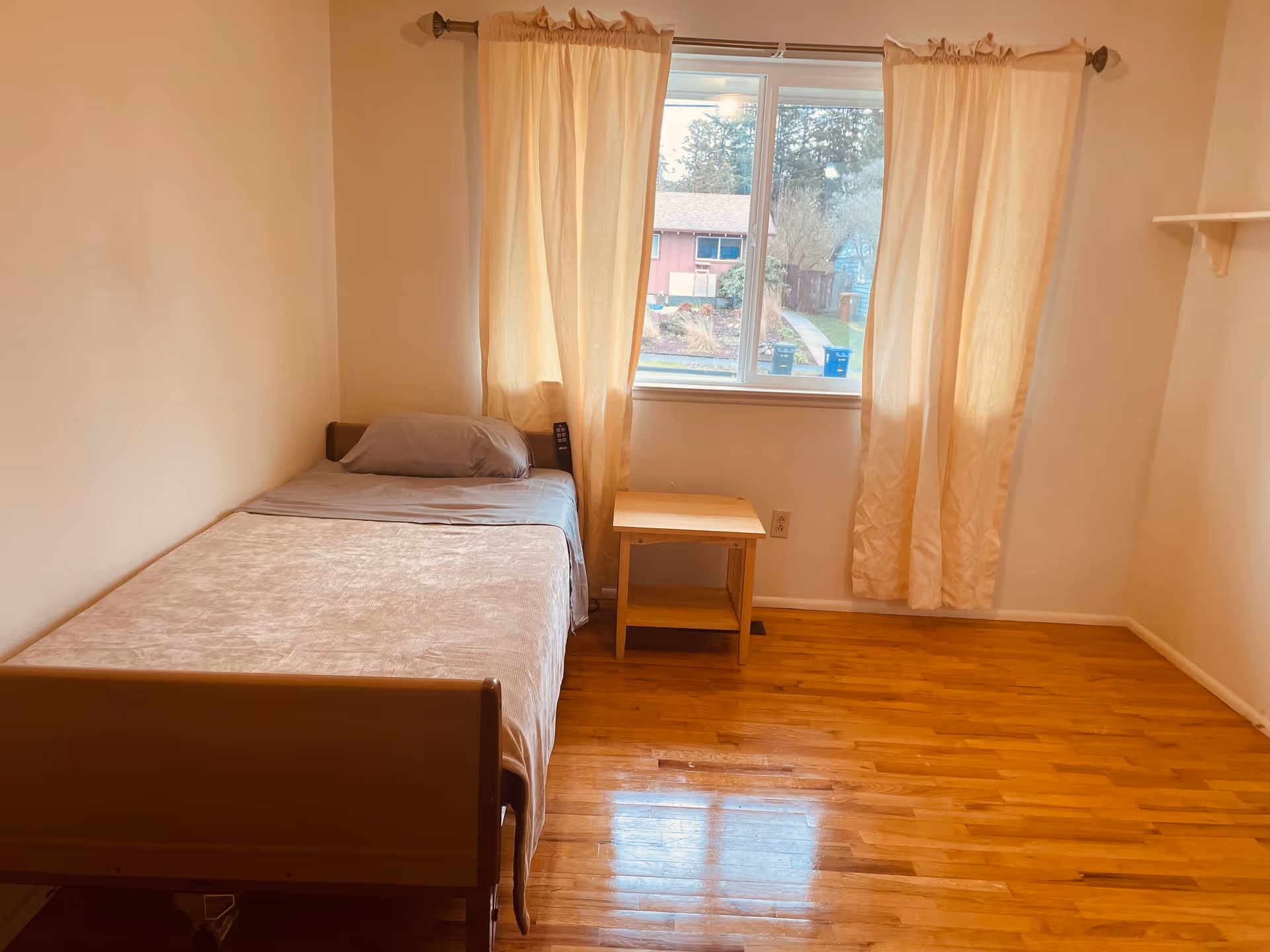 A small bedroom with a single bed, window, light curtains, and wooden floor.