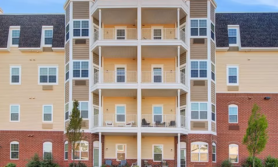 Exterior view of a multi-story senior living facility building with beige siding and red brick on the lower level. The building features multiple balconies with white railings and several windows. There are small trees and shrubs planted in front of the building.