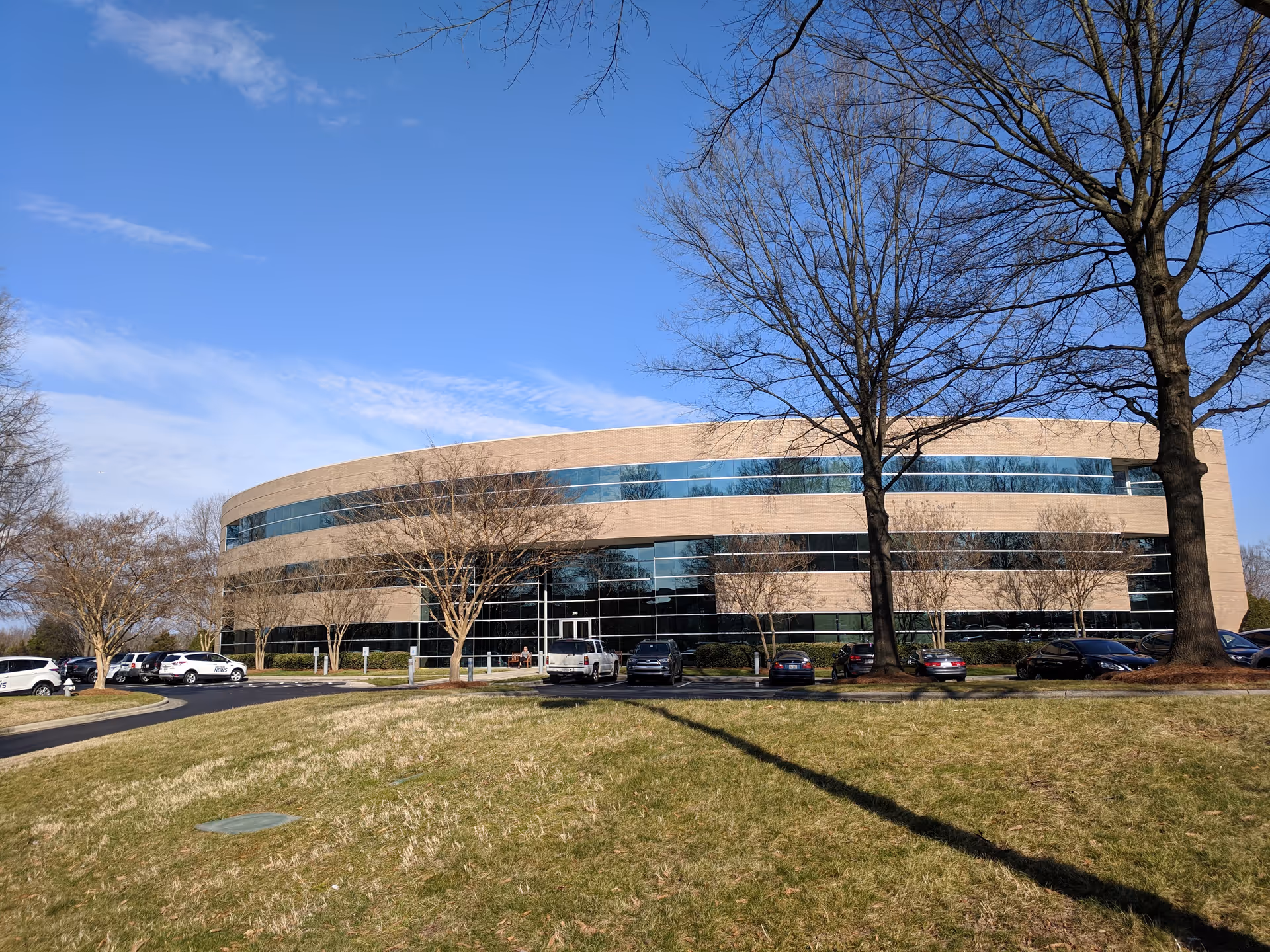 Modern curved glass office building with parking lot and leafless trees.