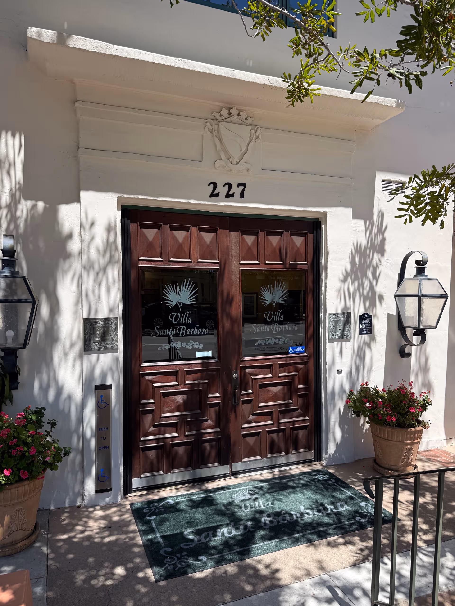 White stucco entrance with dark double doors, Villa Santa Barbara, shaded by trees and potted plants.
