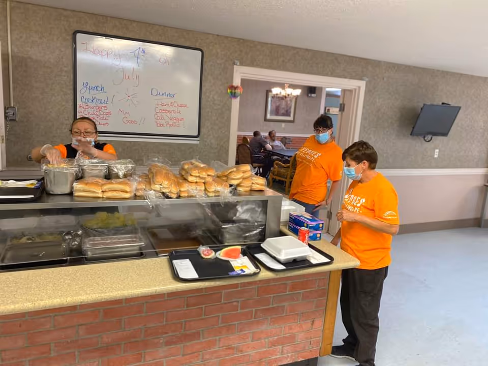Staff in orange shirts serve meals at a brick-front cafeteria counter in a dining room.