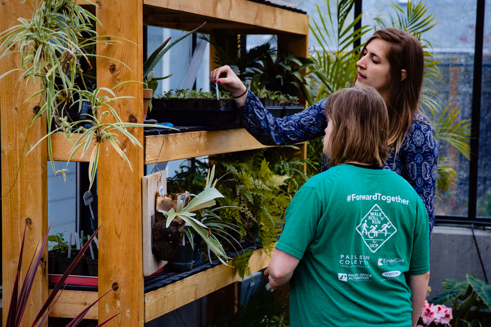 Two women examine potted plants on wooden shelves inside a greenhouse-style indoor garden.