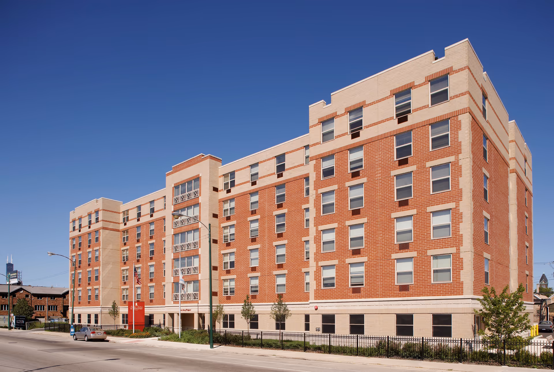 Exterior view of a multi-story brick and beige senior living facility building under a clear blue sky, with a sidewalk, street, and a few parked cars in front.