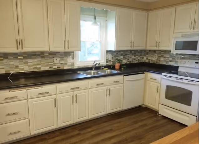 Bright kitchen with white cabinets, dark countertops, sink, stove, and window.