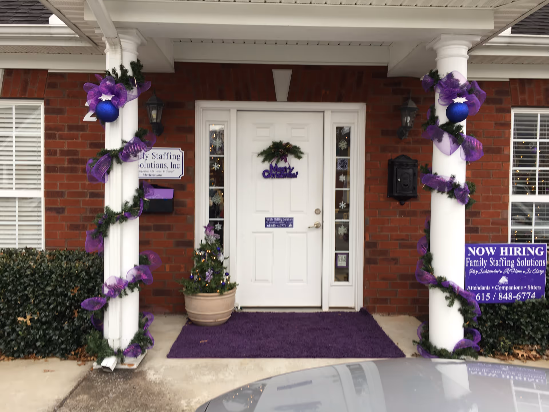 Brick building entrance with white door, purple garlands around columns, wreath, and a 'Now Hiring' sign.