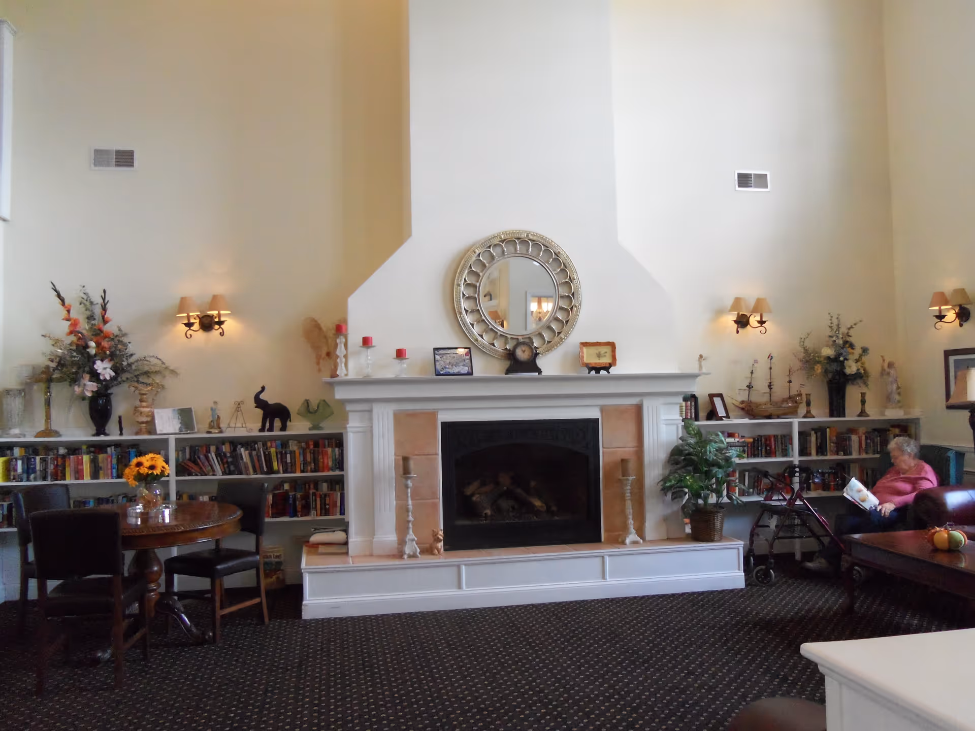 Communal living room with a central fireplace flanked by bookshelves, seating, a round mirror above the mantel, and a resident reading on the right.