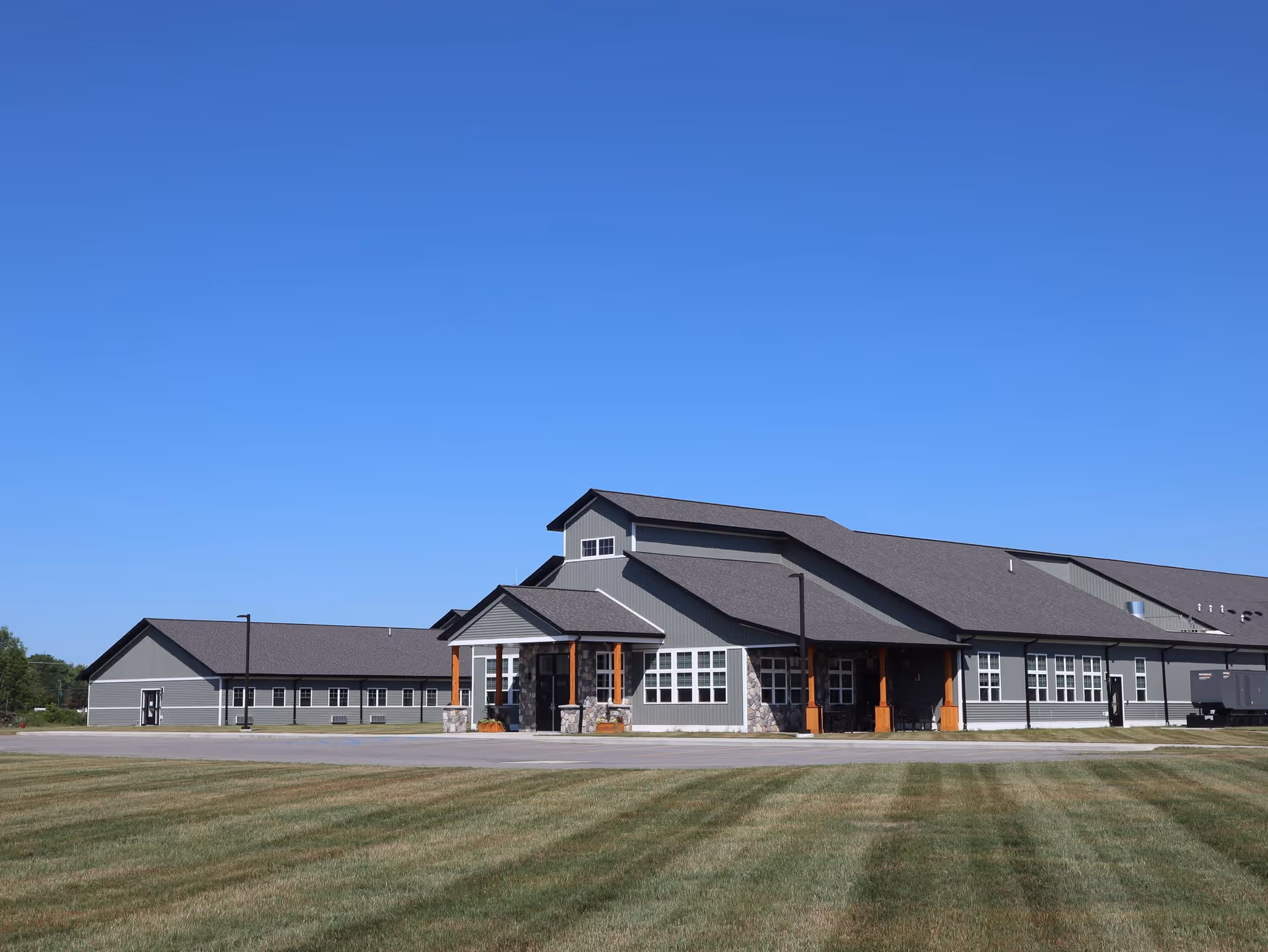 Single-story gray building with gabled roofs and a covered entrance fronting a large grassy lawn under a clear blue sky.