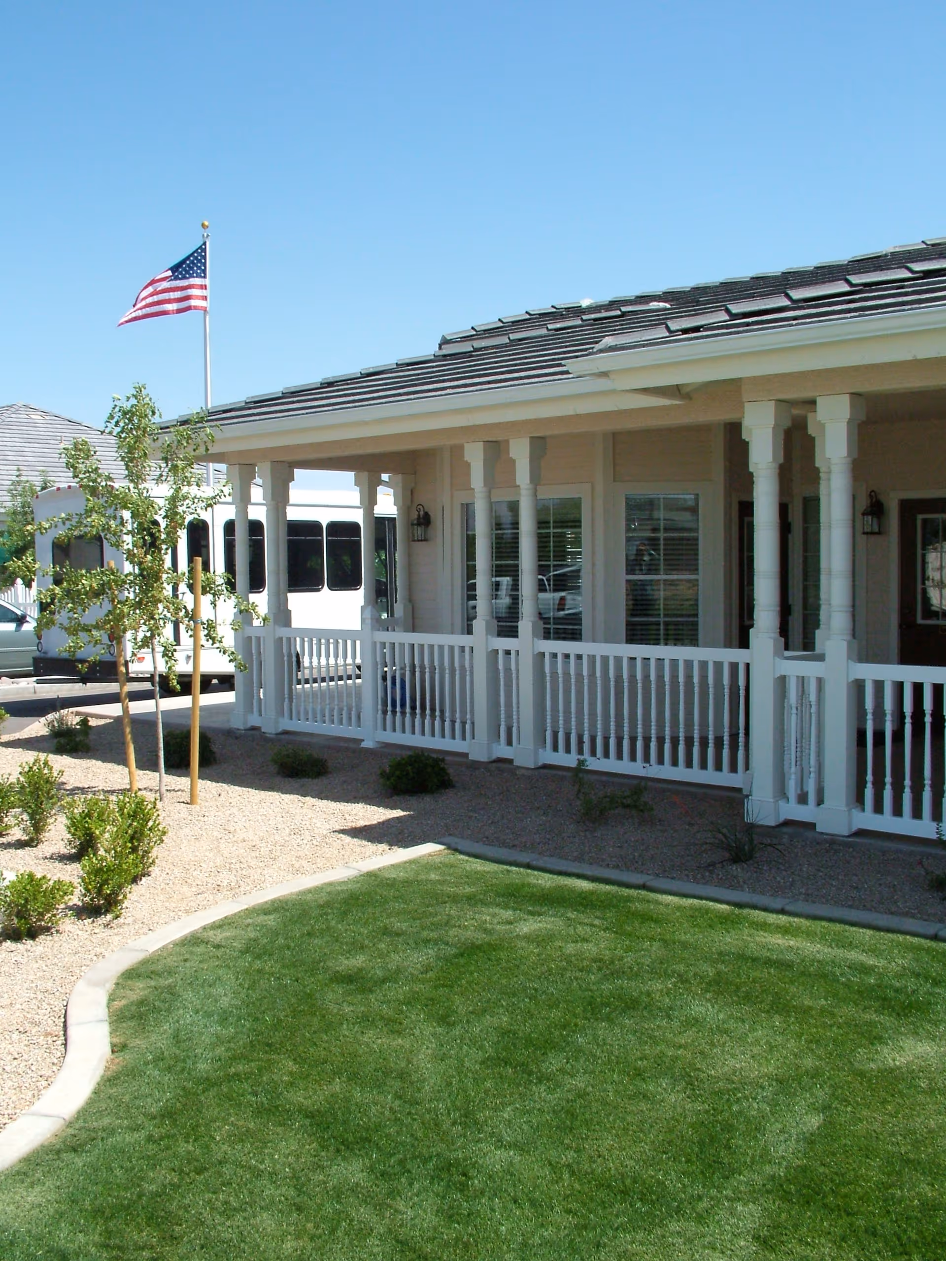Covered porch with white railings and columns in front of a single-story building, a manicured lawn in the foreground and an American flag flying nearby.