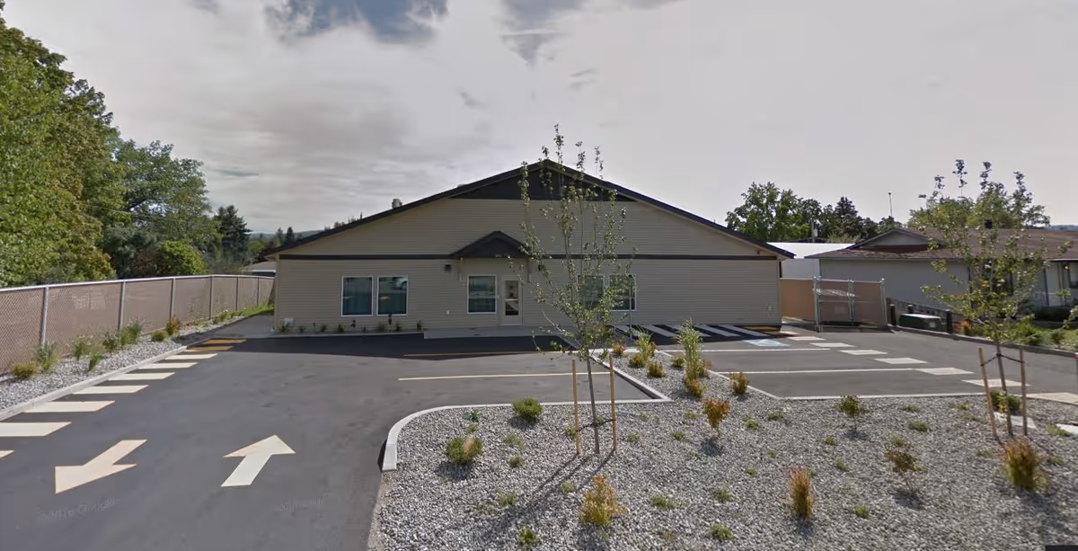Single-story beige building viewed from its parking lot with painted directional arrows, small trees and gravel landscaping beds.