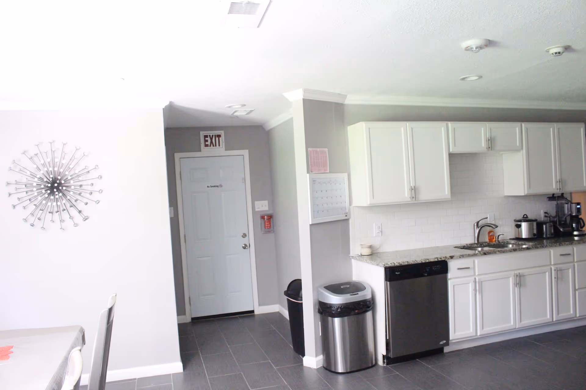 Interior view of a kitchen area in a residential care home featuring white cabinets, a granite countertop, a stainless steel dishwasher, a sink with a faucet, and various kitchen appliances. To the left, there is a dining table with chairs and a decorative wall clock. In the background, there is a white door with an exit sign above it and a fire extinguisher mounted on the wall.