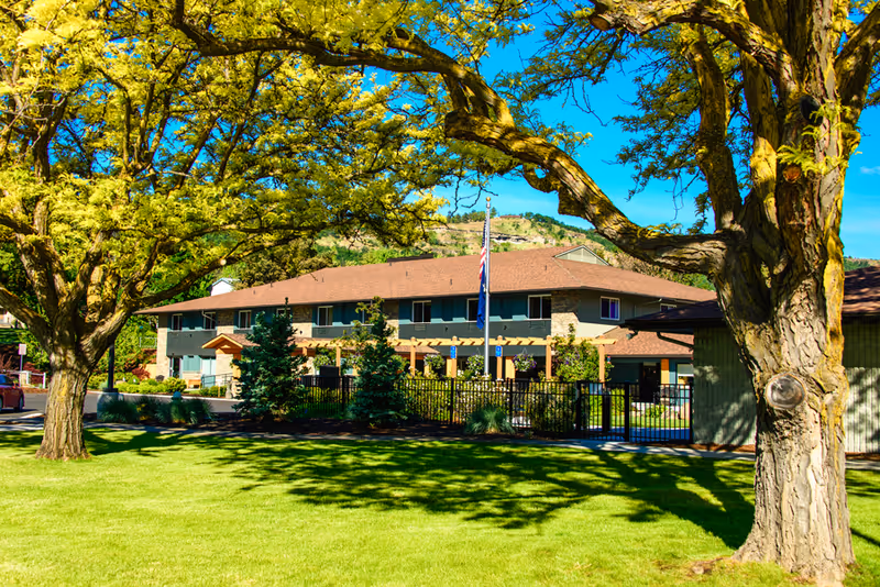 Exterior view of Columbia Basin Care facility with a two-story building surrounded by trees and a well-maintained lawn under a clear blue sky. An American flag is visible in front of the building.