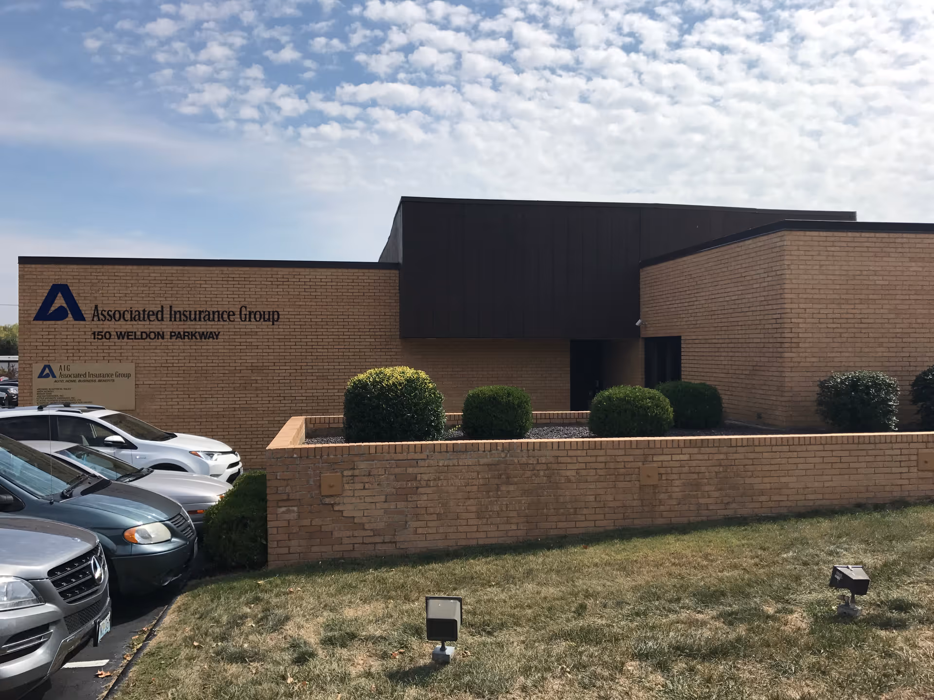 Brick office building front with parked cars and neatly trimmed shrubs.