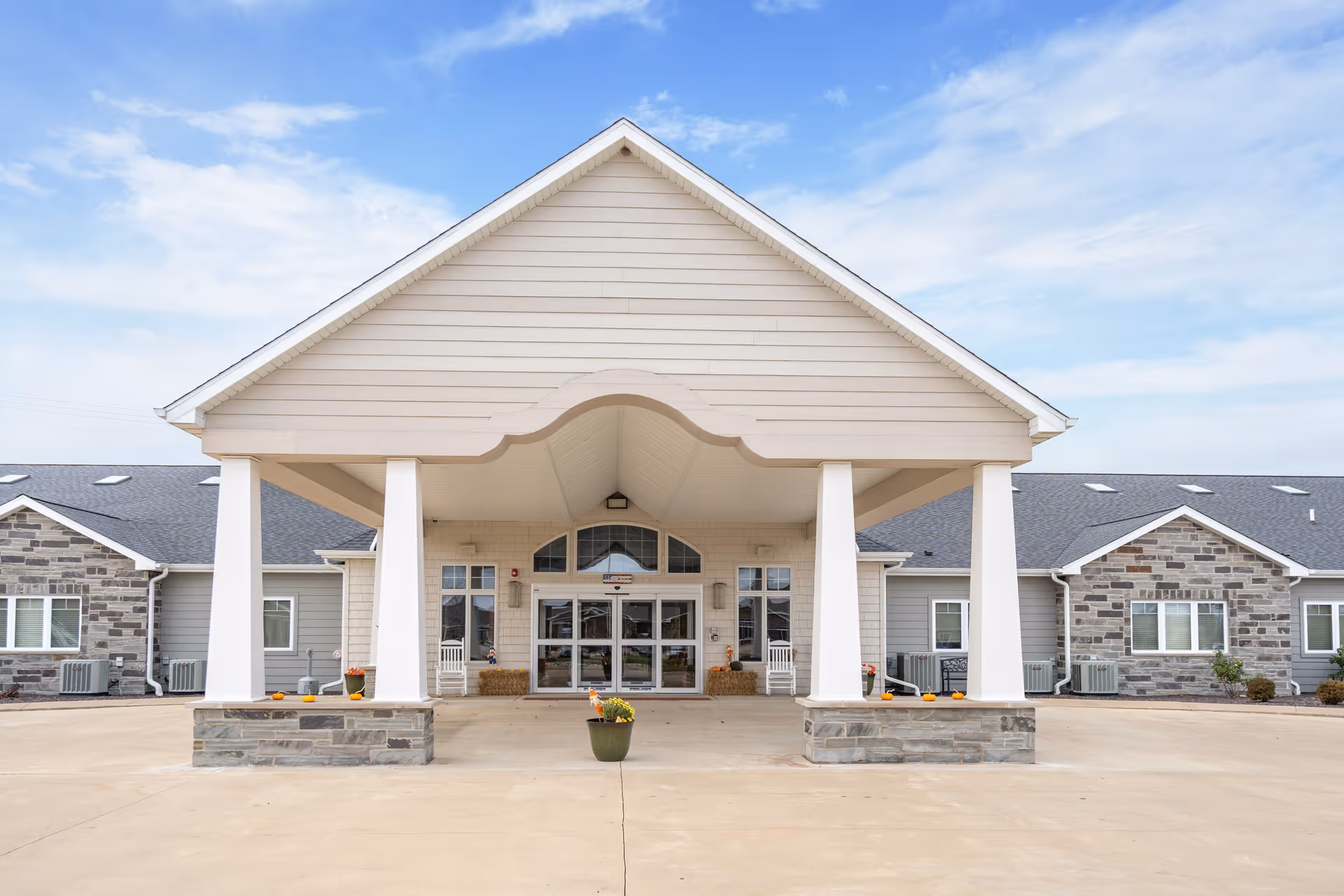 Front exterior view of Reflections Memory Care facility in Washington, IL, showing a large covered entrance with white pillars and stone accents, a concrete driveway, and a partly cloudy sky.