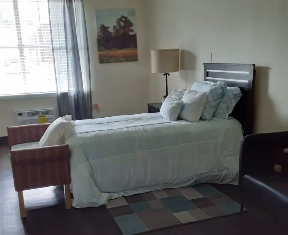 A single bedroom in a senior facility featuring a made bed, lamp, window, and seating.