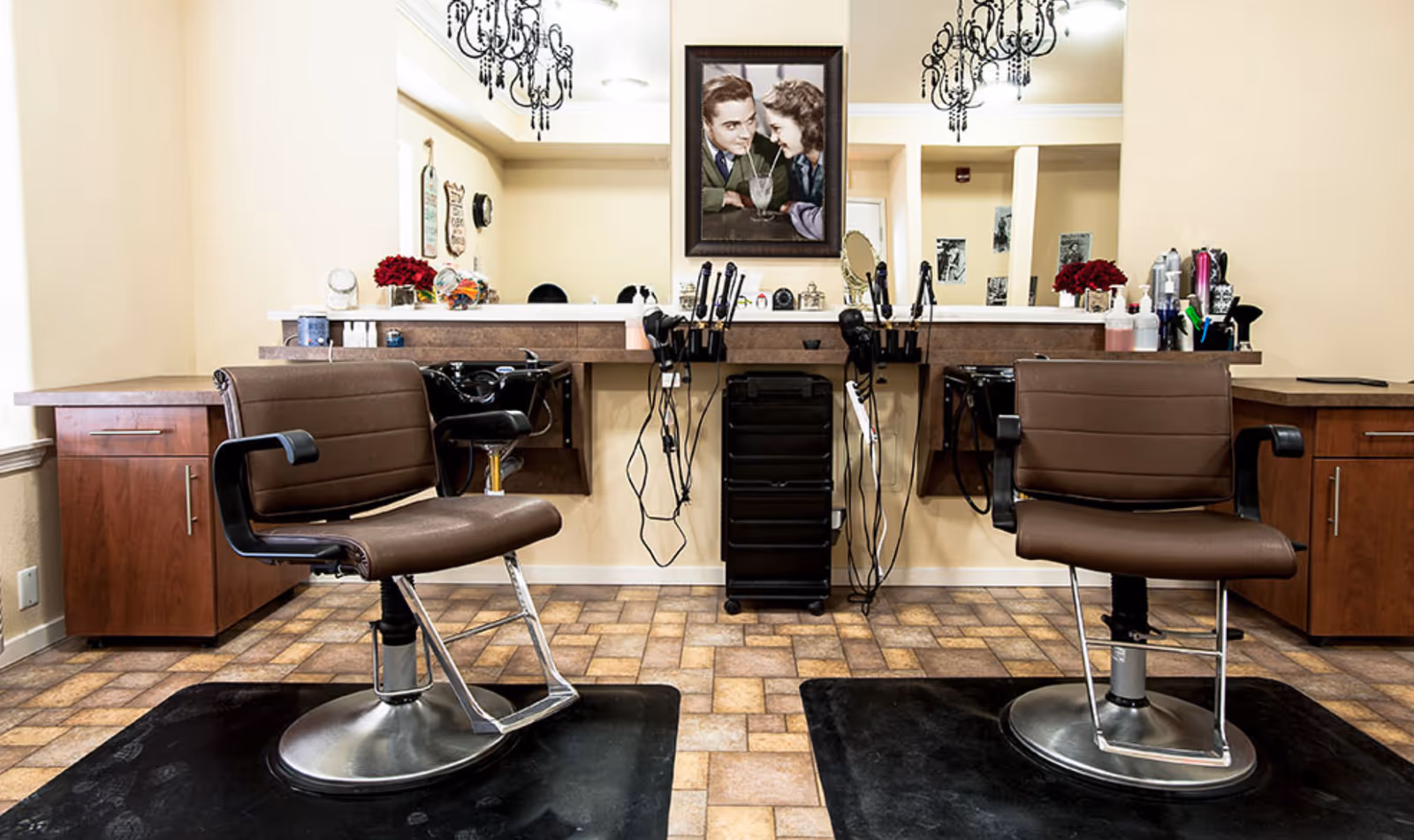 Two brown salon chairs facing a mirrored styling station with hair tools and a framed portrait above.