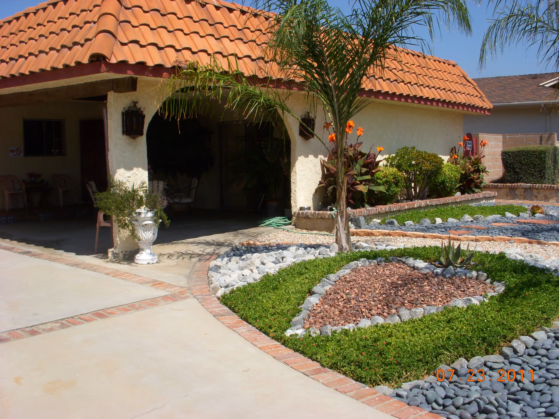 Outdoor view of a facility with a tiled roof and a landscaped garden featuring a palm tree, decorative rocks, and various plants. There is a covered patio area with chairs and potted plants.
