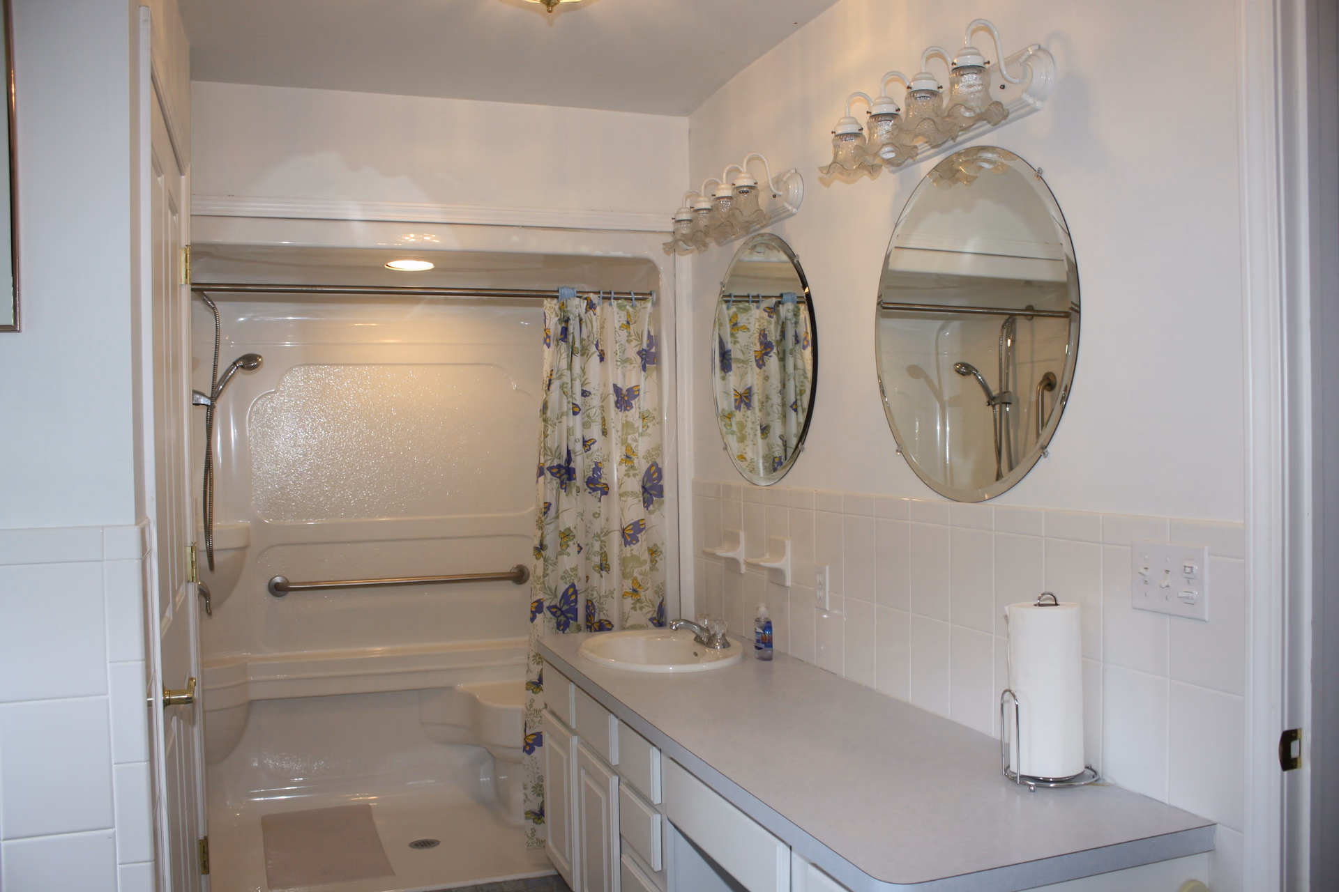 Bathroom with a shower-tub with grab bar and floral curtain, dual oval mirrors, and a long sink vanity.