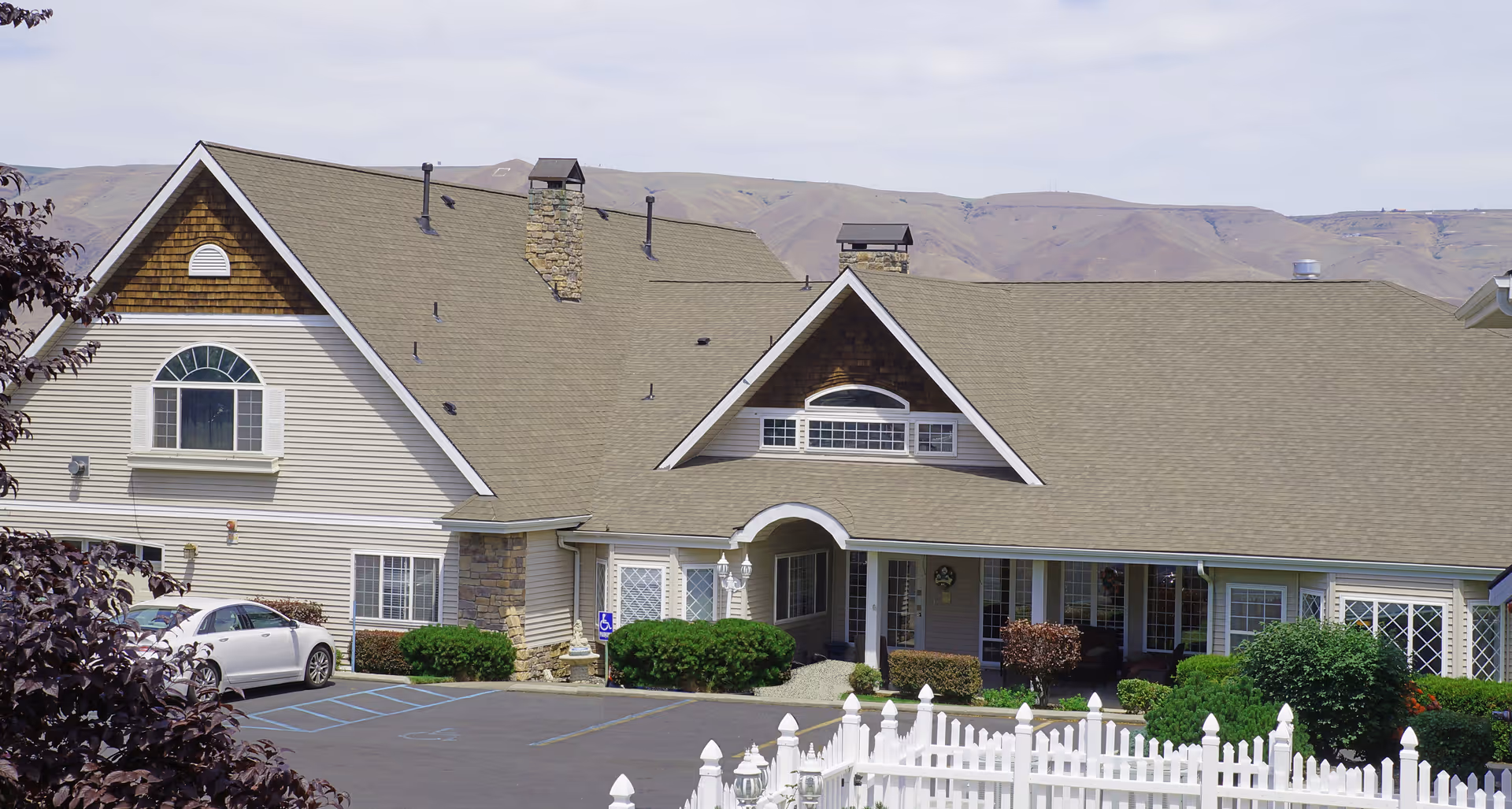 Front exterior of a beige senior living building with pitched roofs, a white picket fence, parked cars, and hills in the background.