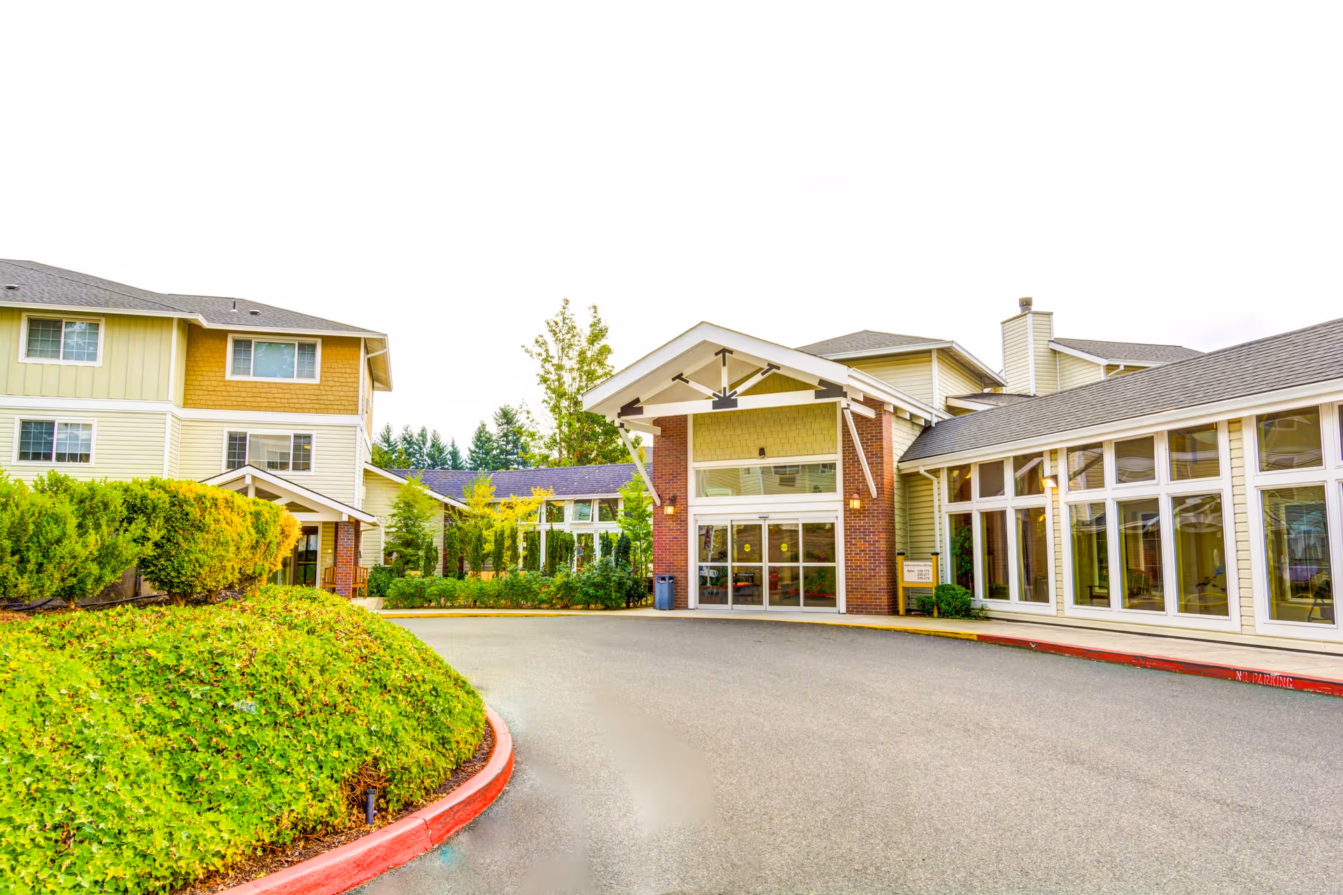 Front exterior of a senior living facility showing a covered entrance, large windows, and a landscaped driveway.