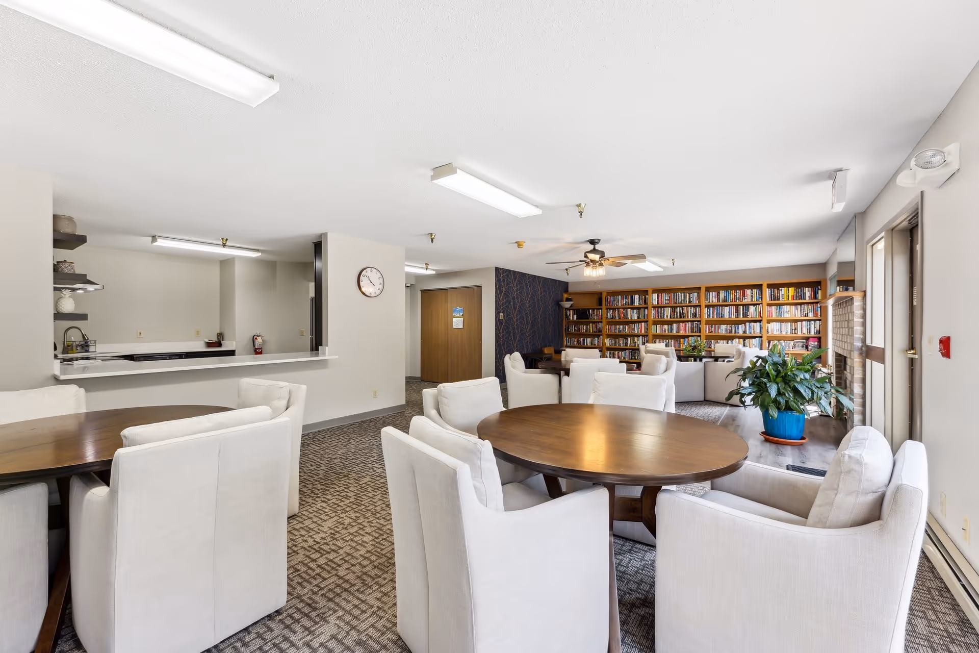 A bright and spacious common area with round wooden tables surrounded by white cushioned chairs. In the background, there is a large bookshelf filled with books, a ceiling fan with lights, and a cozy seating area with armchairs. To the left, there is a kitchen counter with shelves holding decorative items. A large potted plant is near the glass door on the right side, and the room has carpeted flooring and fluorescent ceiling lights.