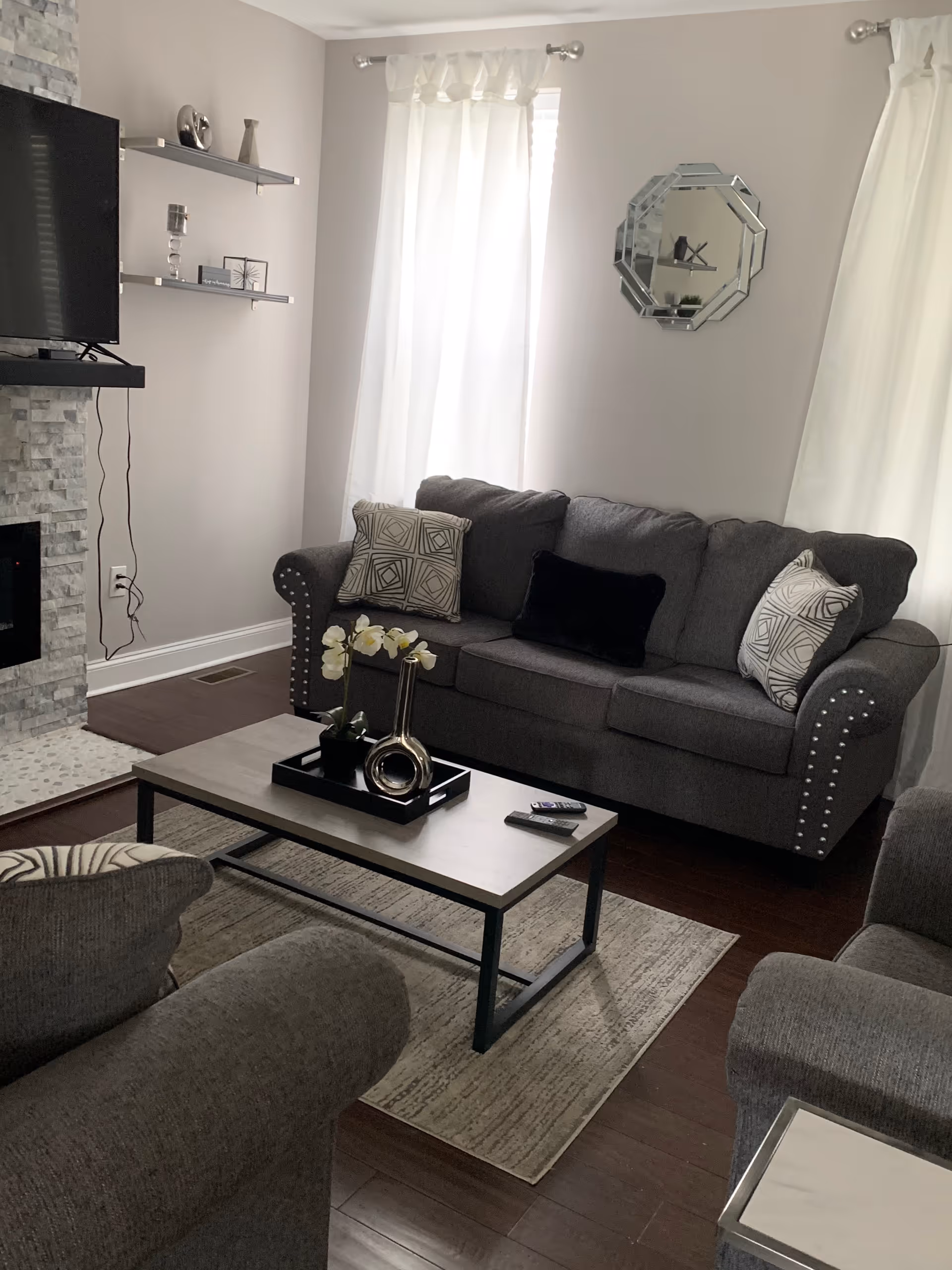 Cozy gray living room with sofa, coffee table, fireplace, TV, shelves, curtains, and large windows.