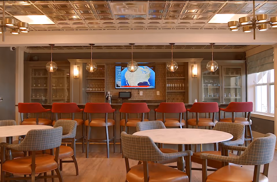 A dining/lounge area with round marble-top tables and upholstered chairs facing a bar with red stools, pendant lights, and a wall-mounted TV.