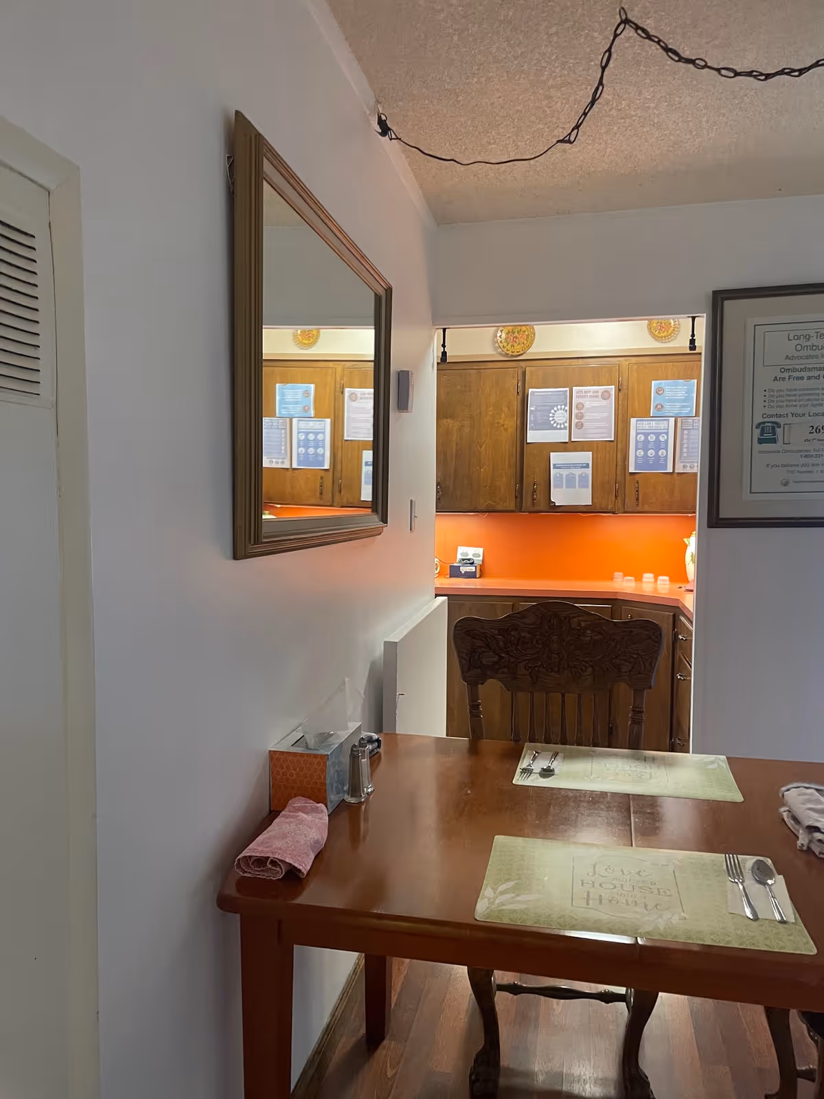 A dining area with a wooden table set for two with placemats, utensils, a tissue box, and a folded cloth napkin. A wooden chair with carved details is positioned at the table. A mirror hangs on the wall reflecting part of the kitchen area, which has wooden cabinets and an orange backsplash. Various notices and papers are posted on the kitchen cabinets.