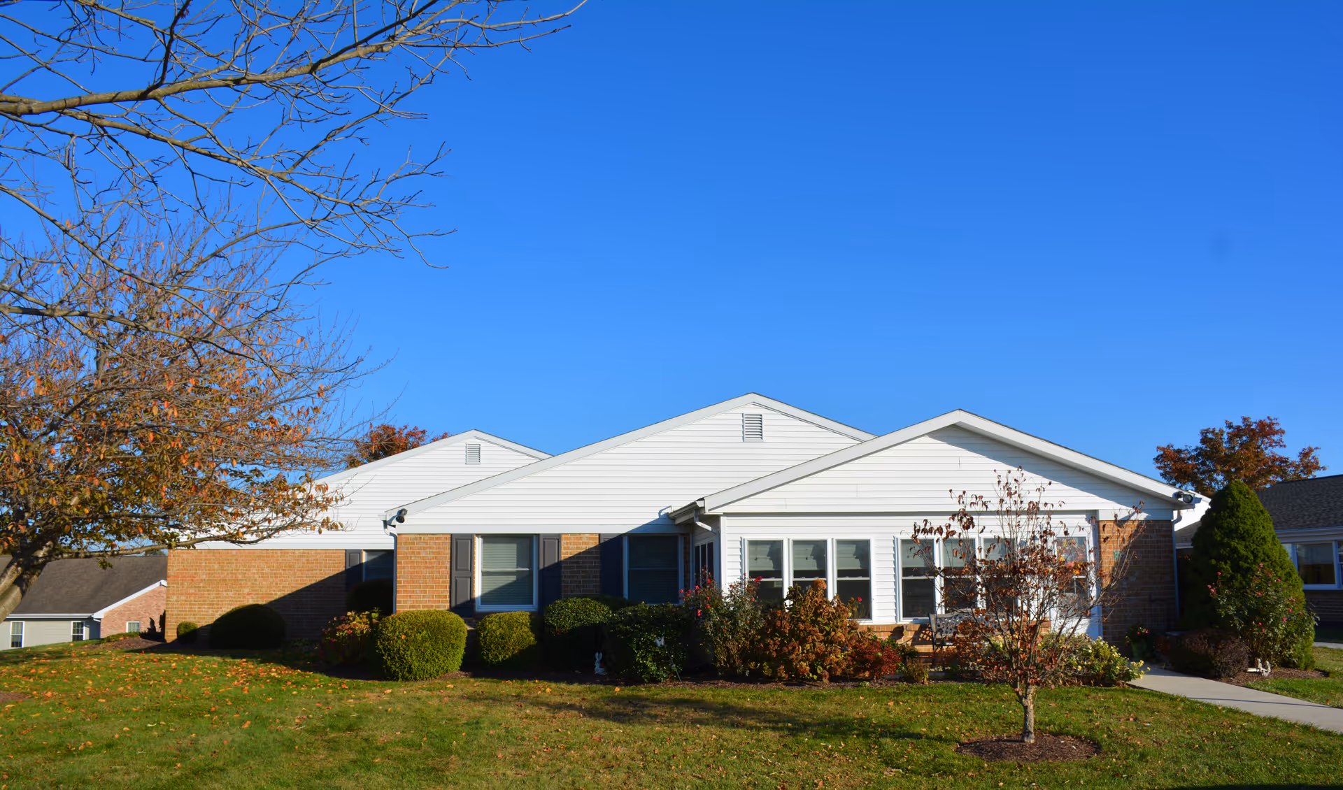Exterior view of a single-story residential building with white siding and brick walls, surrounded by a well-maintained lawn, bushes, and small trees under a clear blue sky.