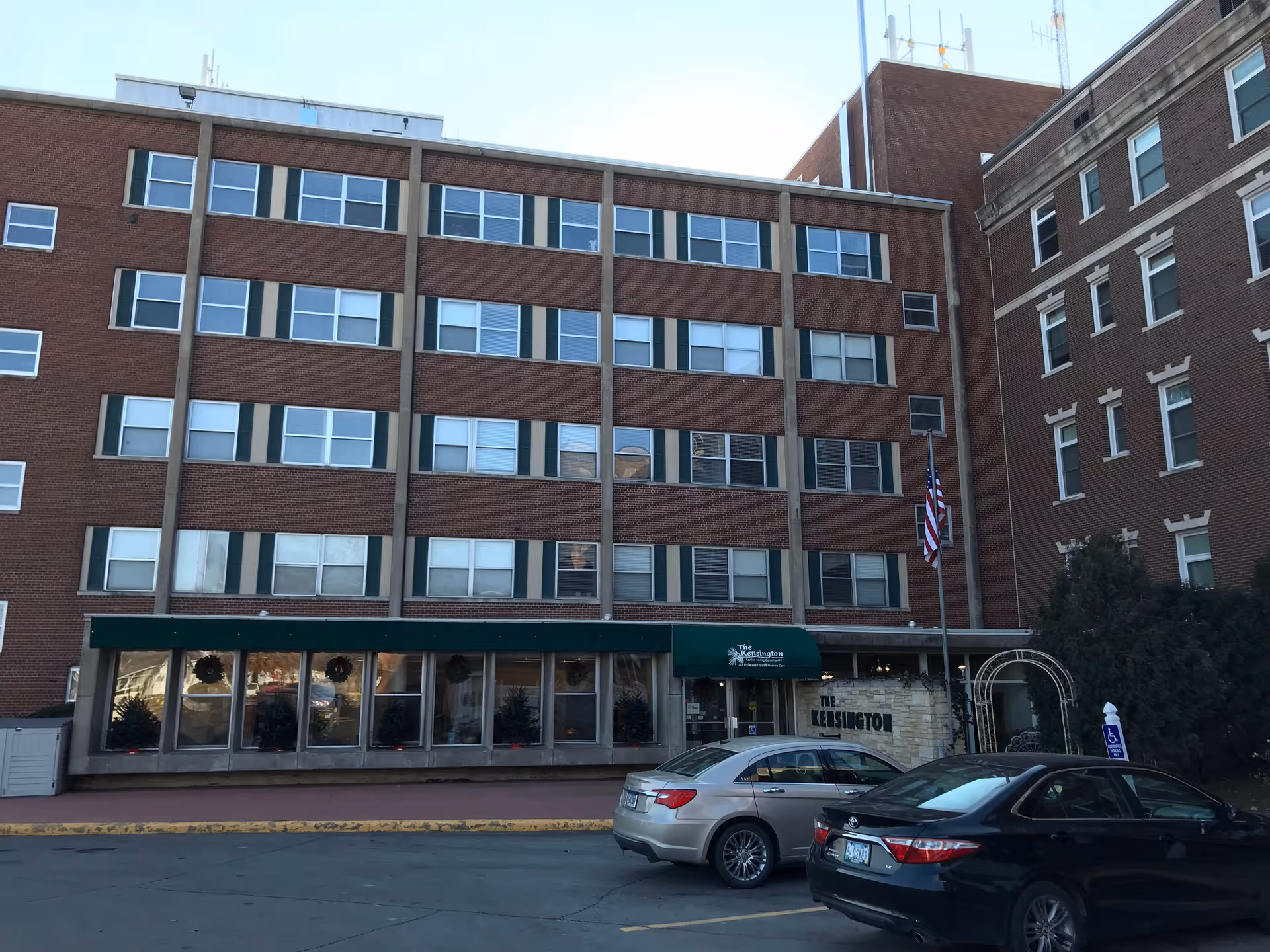 Exterior view of The Kensington senior living facility showing a multi-story brick building with many windows, a green awning over the entrance, an American flag on a flagpole, and two parked cars in front.