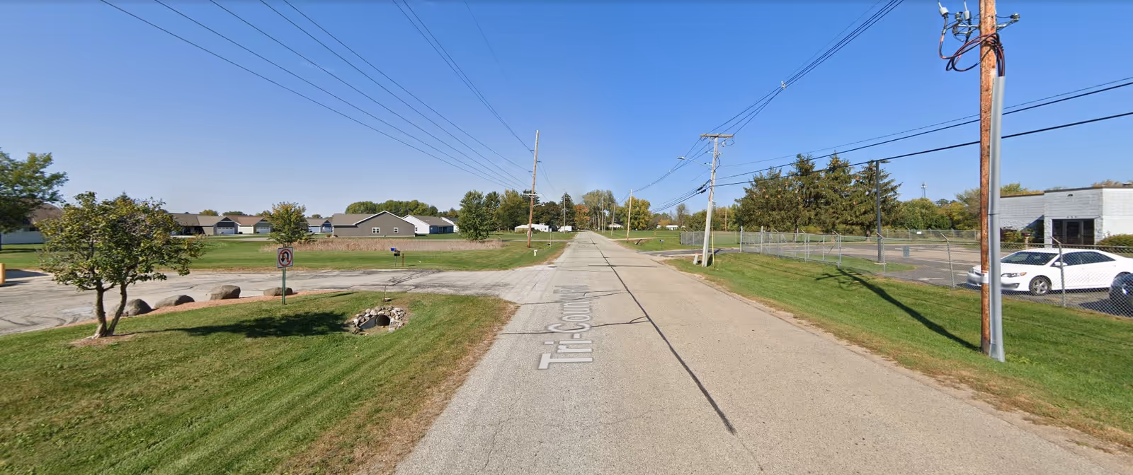 A quiet suburban street with green lawns, utility poles, and power lines running along both sides. There are single-story houses in the distance on the left and a fenced parking lot with a white car on the right under a clear blue sky.