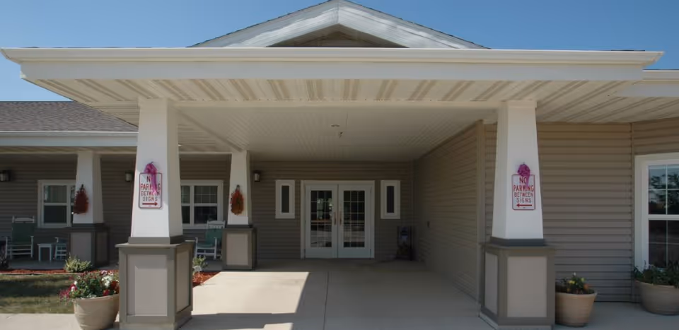 Entrance of a building with a covered porch supported by four white columns. The building has beige siding and white trim, with double glass doors in the center. There are potted plants on either side of the entrance and 'No Parking Between Signs' notices attached to two of the columns. The sky is clear and blue.