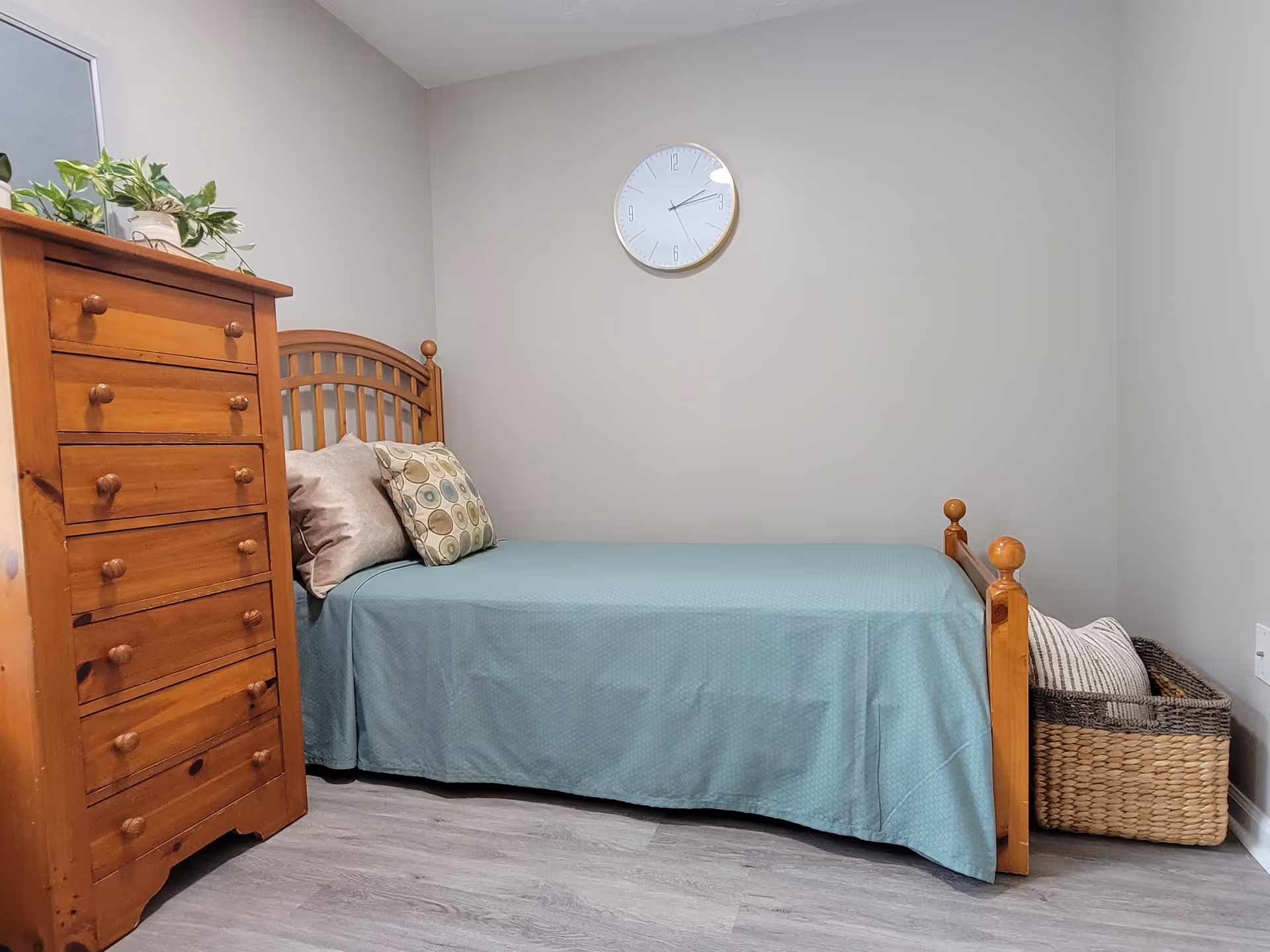 Senior living facility bedroom with wooden bed, dresser, clock, and wicker basket.